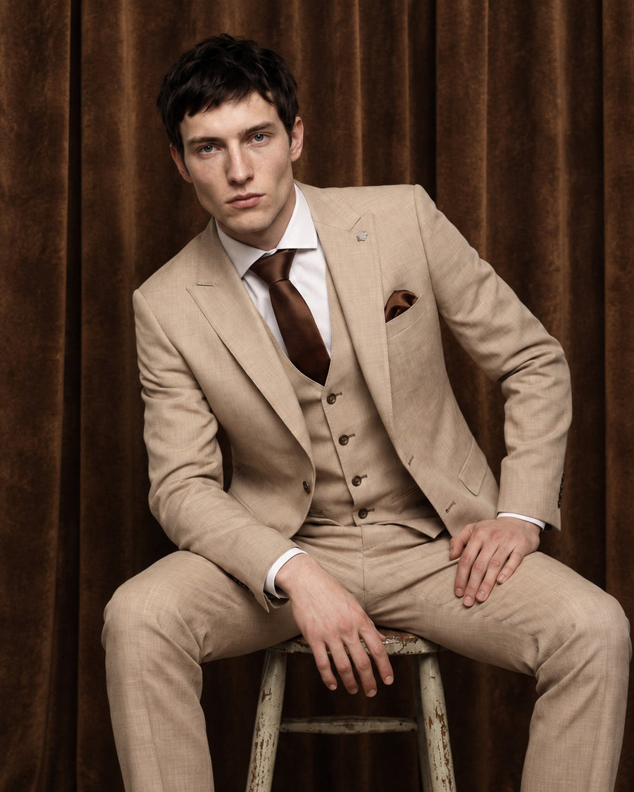 A man wearing a beige three-piece suit with a white shirt, brown tie, and brown pocket square, sitting on a rustic wooden stool against a plain off-white background.