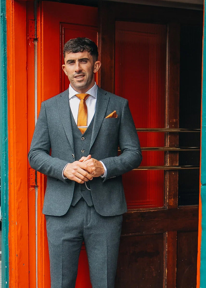 Aaron Gillane wearing a grey three-piece suit, white dress shirt, and orange tie stands in a doorway with a bright orange frame and teal wall.