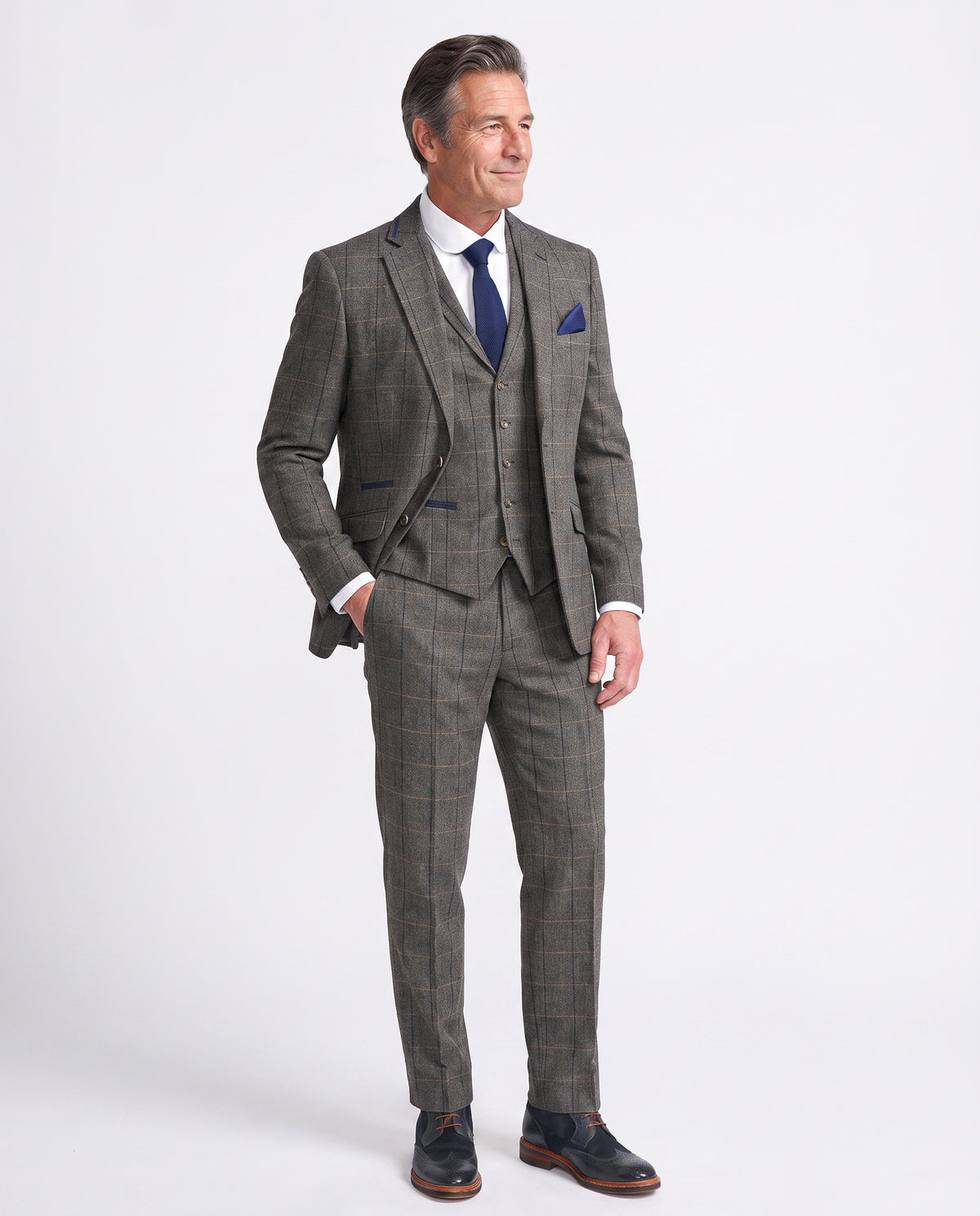 A man modeling a three-piece grey windowpane plaid suit with a white dress shirt, blue tie, and blue pocket square, standing against a light grey studio background.