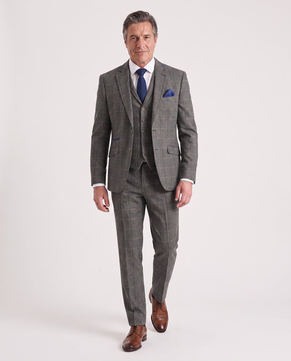 A man wearing a gray three-piece windowpane plaid suit with a white dress shirt, blue tie, and blue pocket square, walking against a light gray studio background.