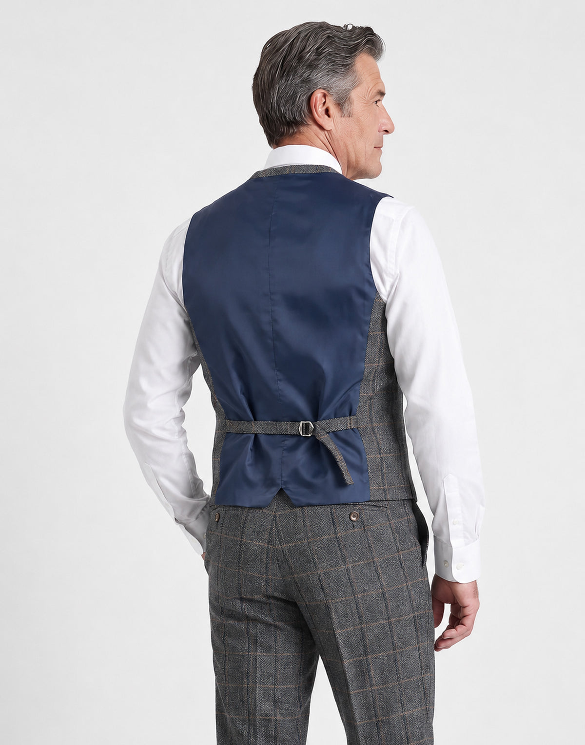 A man shown from the back wearing a navy blue waistcoat over a gray and brown windowpane plaid suit, paired with a white dress shirt, against a light gray studio background.
