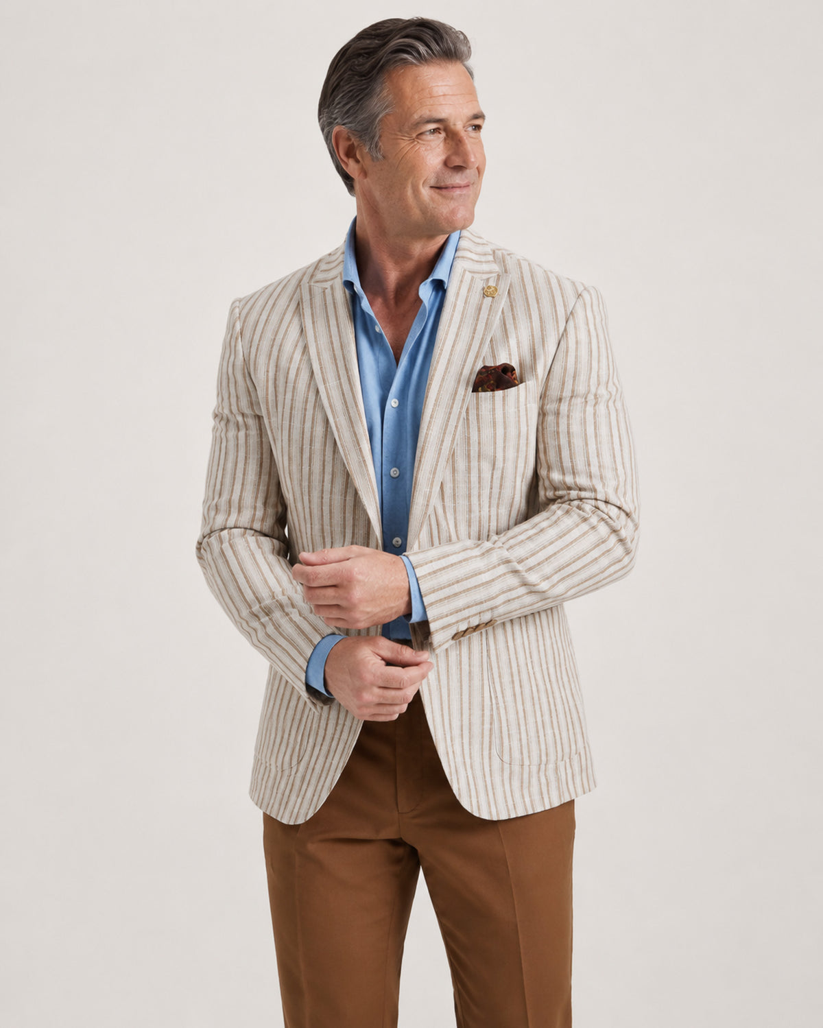 A man wearing a beige and white vertically striped blazer over a light blue dress shirt, paired with brown trousers, photographed against a plain light gray studio background.