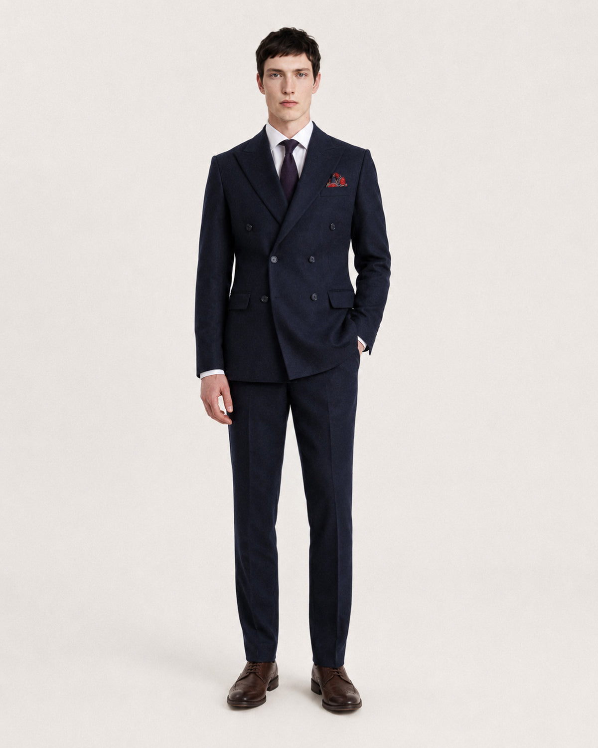 A man modeling a dark navy double-breasted suit with a white dress shirt, a dark patterned tie, a red pocket square, and brown leather dress shoes, standing against a plain light background.