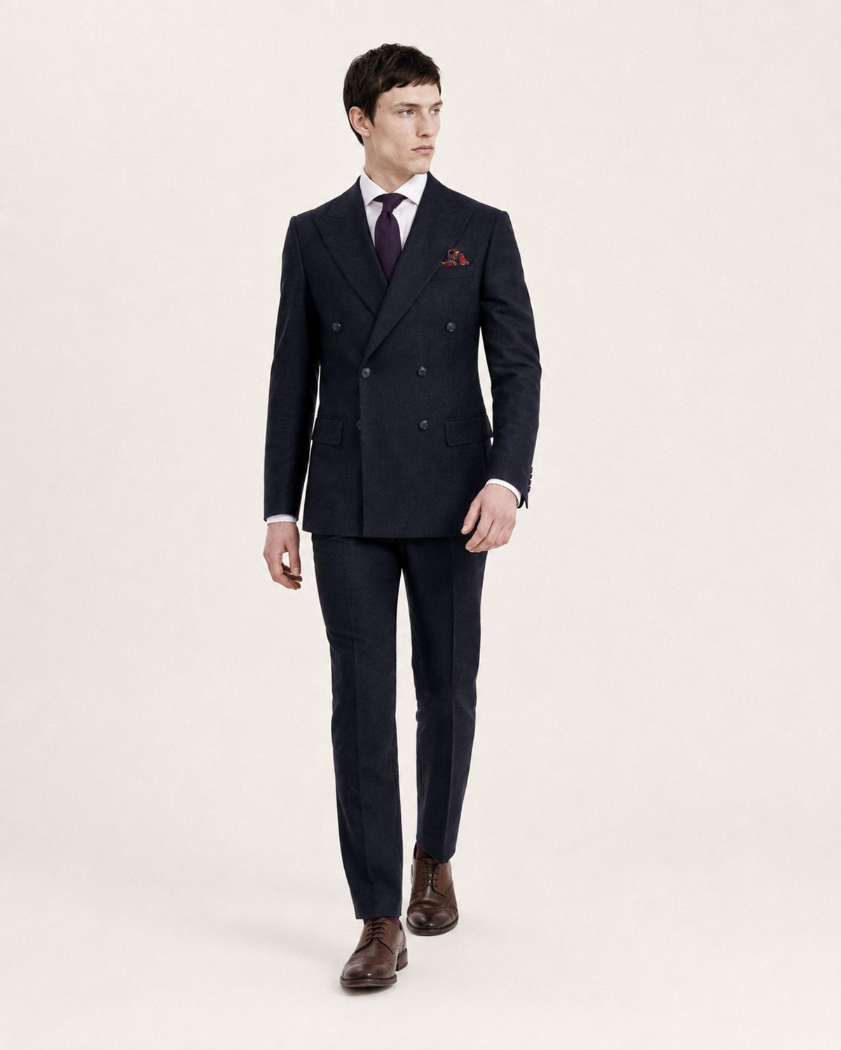 A man modeling a dark navy double-breasted suit with a white dress shirt, a dark purple tie, and a red pocket square, standing against a plain off-white background.