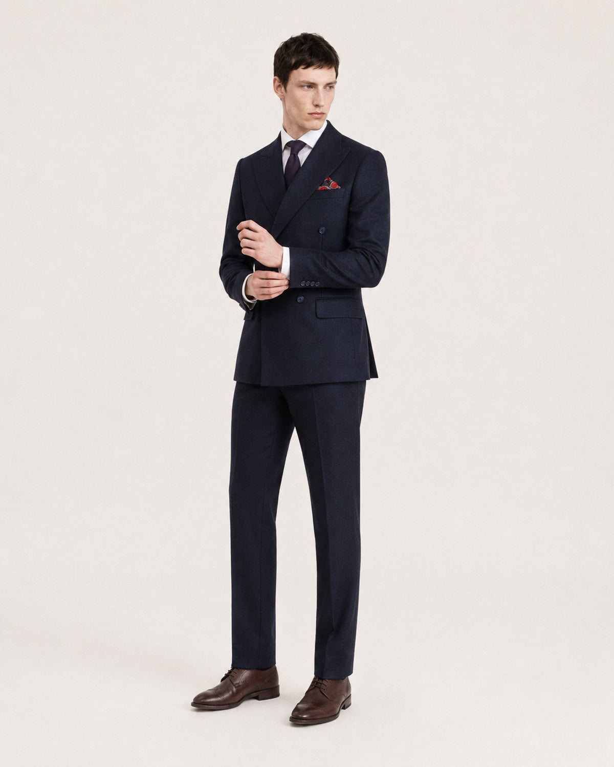 A man modeling a dark navy double-breasted suit with a white dress shirt, dark tie, and a red pocket square, standing against a plain light background.