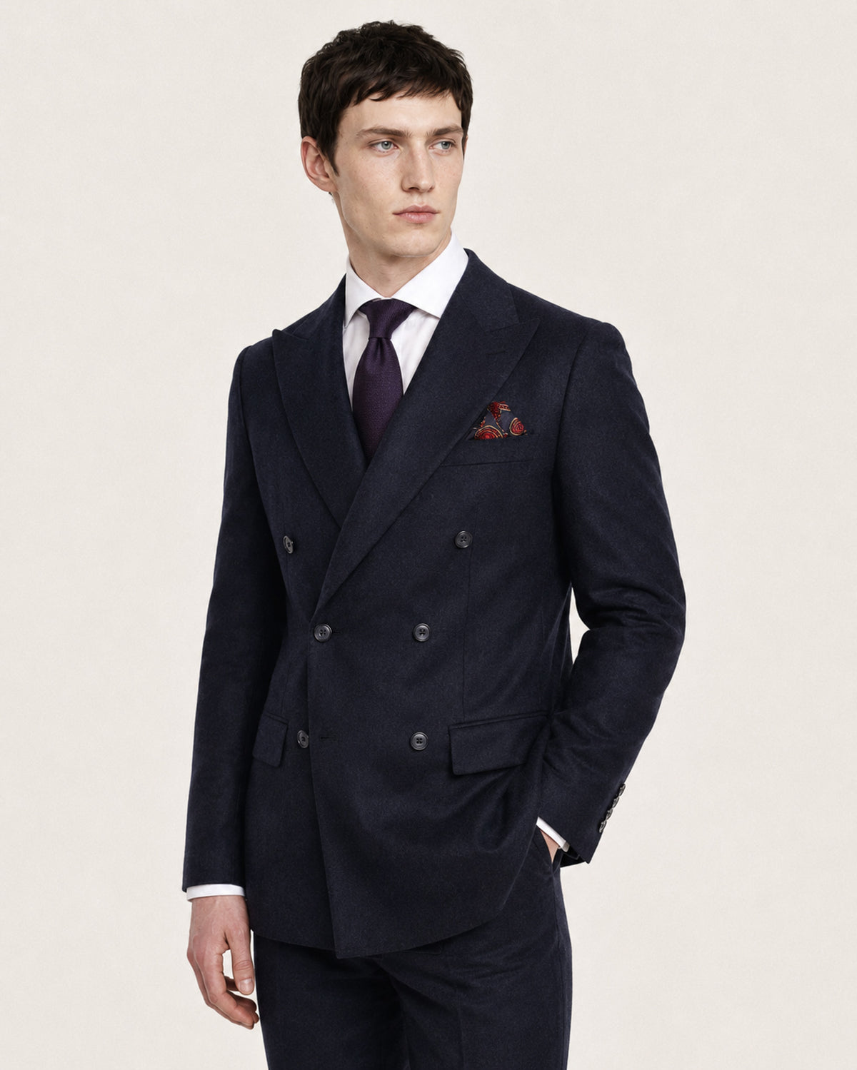 A man wearing a dark navy double-breasted suit with a white dress shirt, a dark purple tie, and a red patterned pocket square, photographed against a plain light beige studio background.