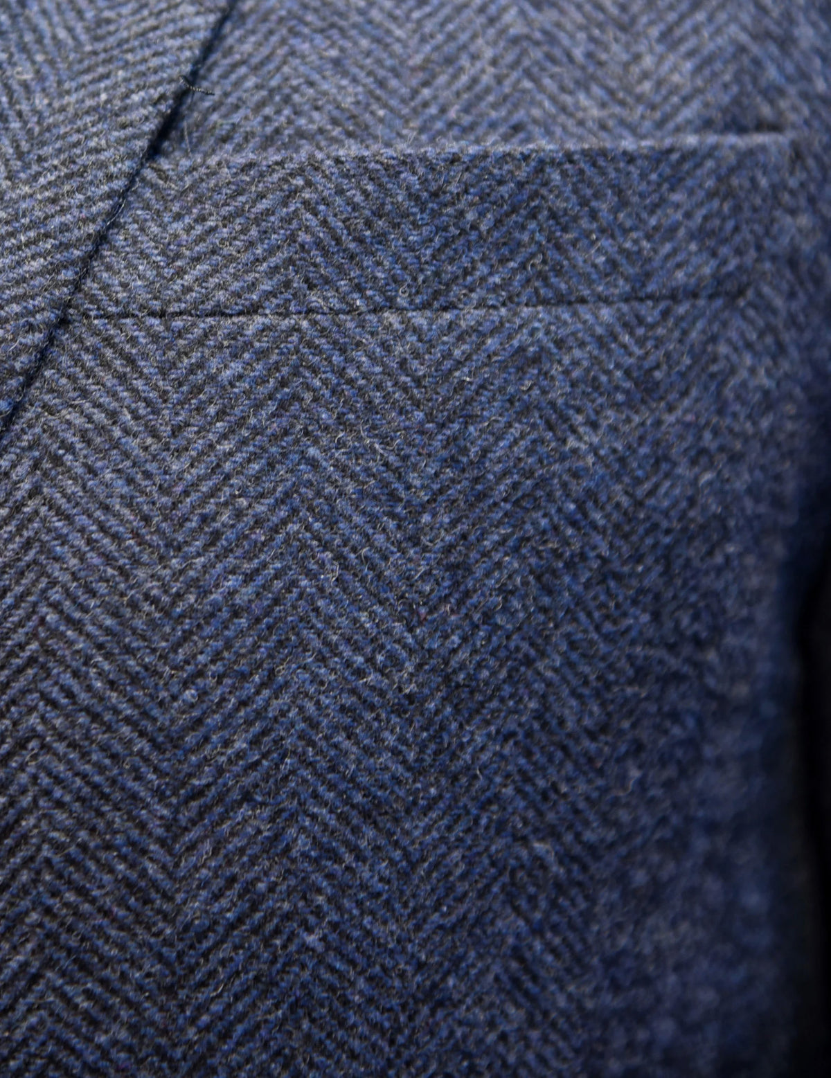 Close-up of a blue and gray herringbone tweed fabric texture, showing the woven pattern and stitched pocket detail.