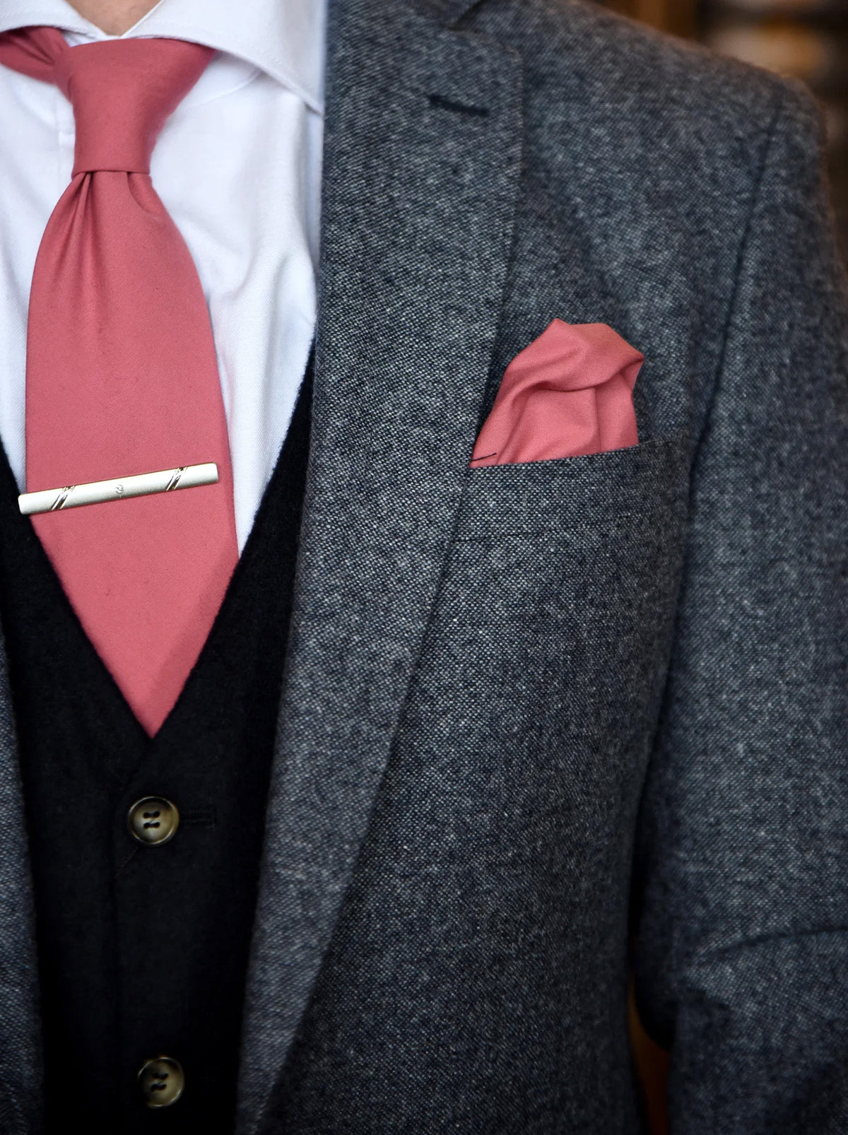 A close-up of a men's formal outfit featuring a textured gray tweed suit jacket, a black wool vest, a white dress shirt, a dusty rose pink tie with a silver tie bar, and a matching dusty rose pocket square.