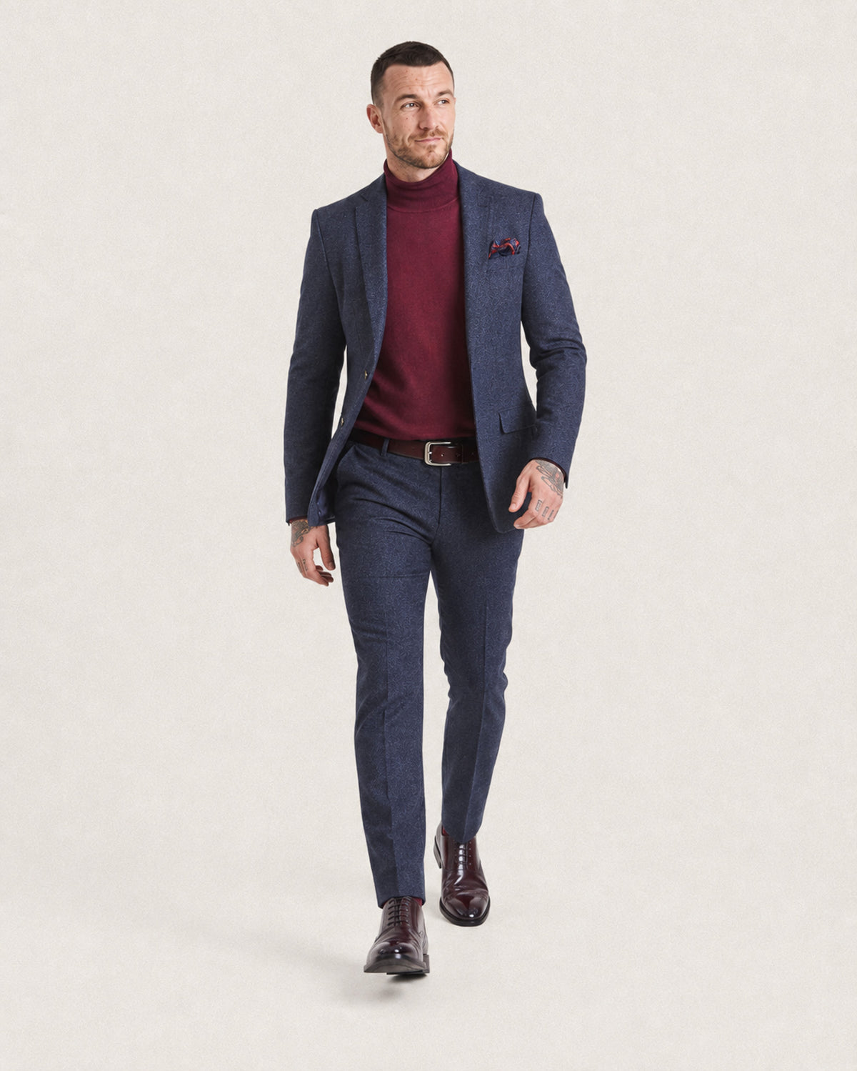 A man modeling a navy blue textured blazer and matching trousers, styled with a burgundy turtleneck sweater, brown leather belt, and dark brown dress shoes, against a light beige studio background.