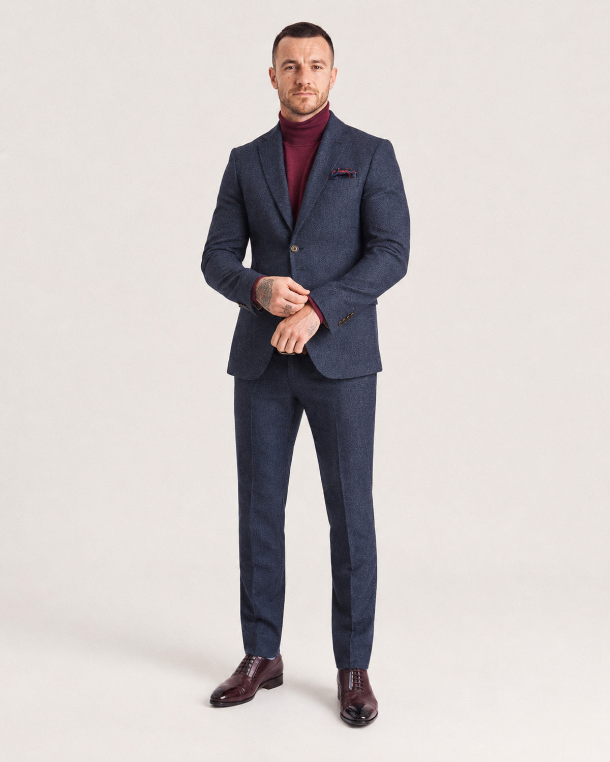 A man modeling a navy blue two-piece suit with a subtle textured pattern, worn over a burgundy turtleneck sweater and paired with dark brown dress shoes, against a plain light gray studio background.