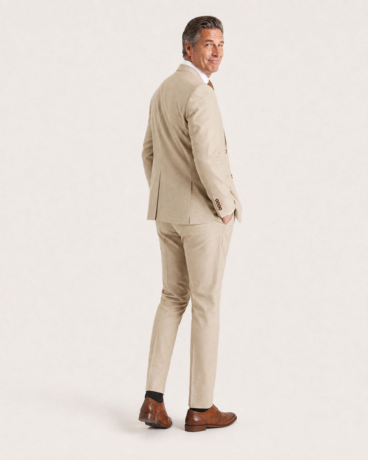 A man wearing a light beige two-piece suit, white dress shirt, and brown leather dress shoes, shown from a side-back angle against a plain white studio background.