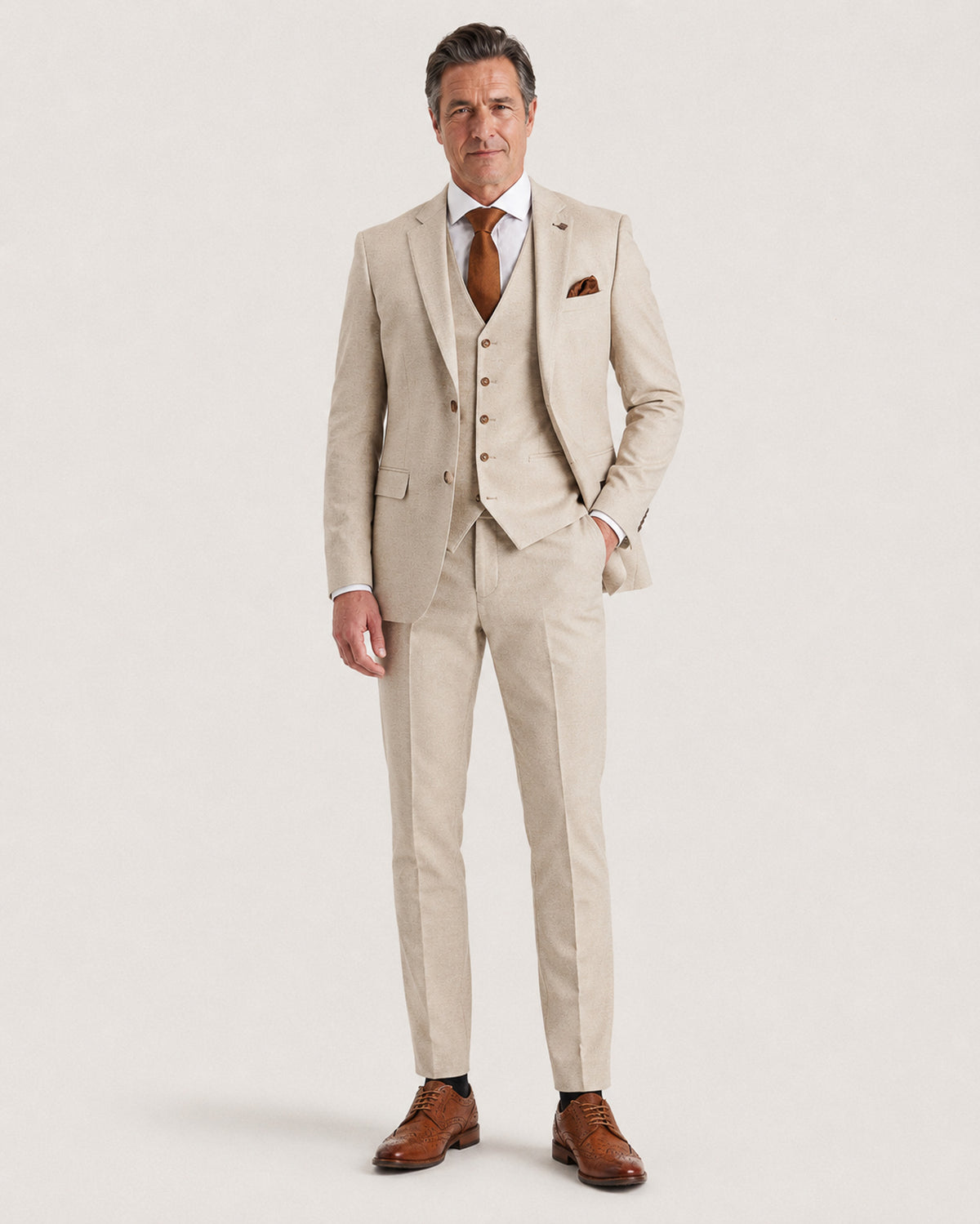 A man modeling a light beige three-piece suit with a matching vest and trousers, styled with a white dress shirt, a brown patterned tie, a brown pocket square, and brown leather dress shoes, against a solid off-white background.