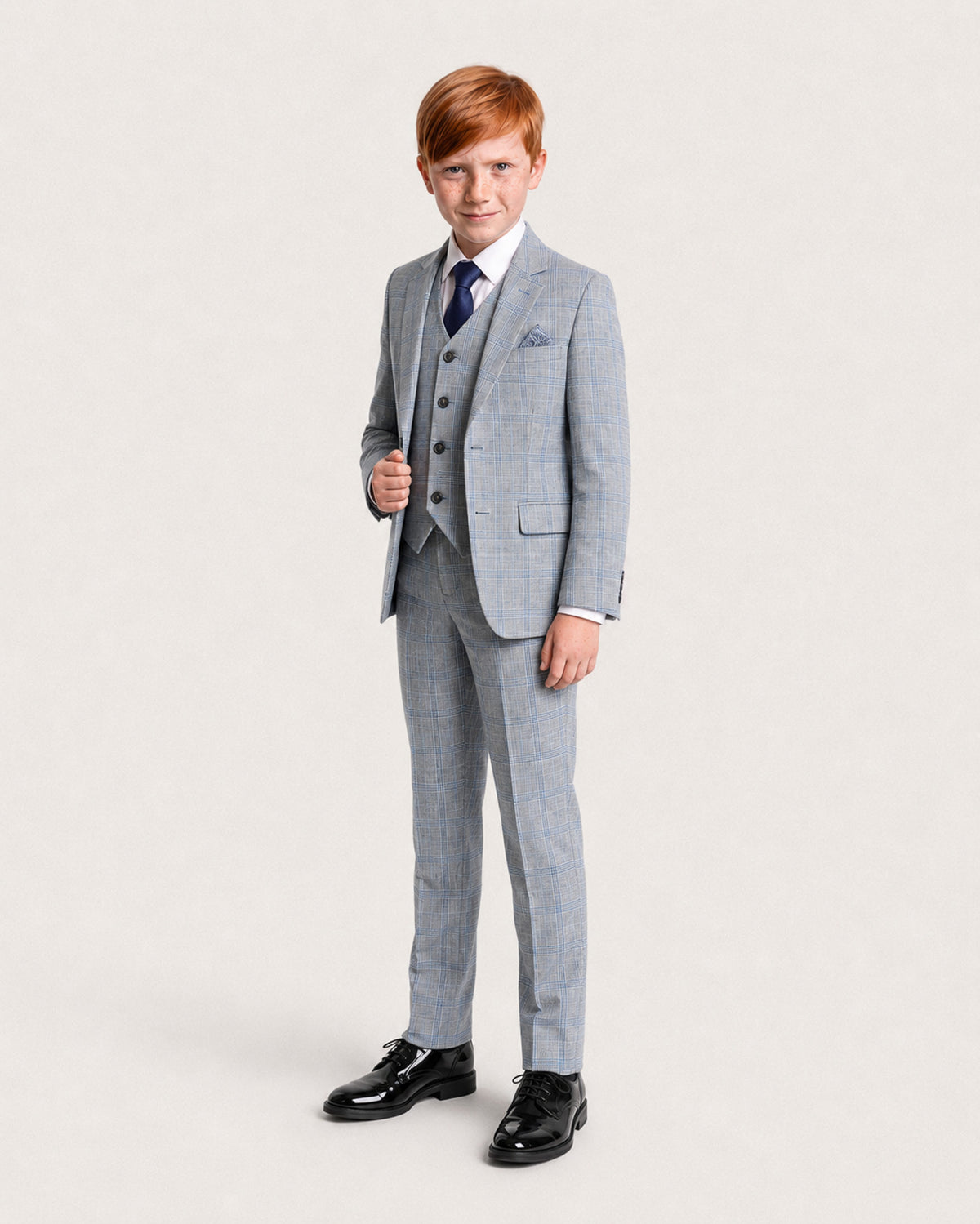A young boy with red hair wearing a light blue and white check three-piece suit, white shirt, blue patterned tie, and black dress shoes, standing against a light gray studio background.