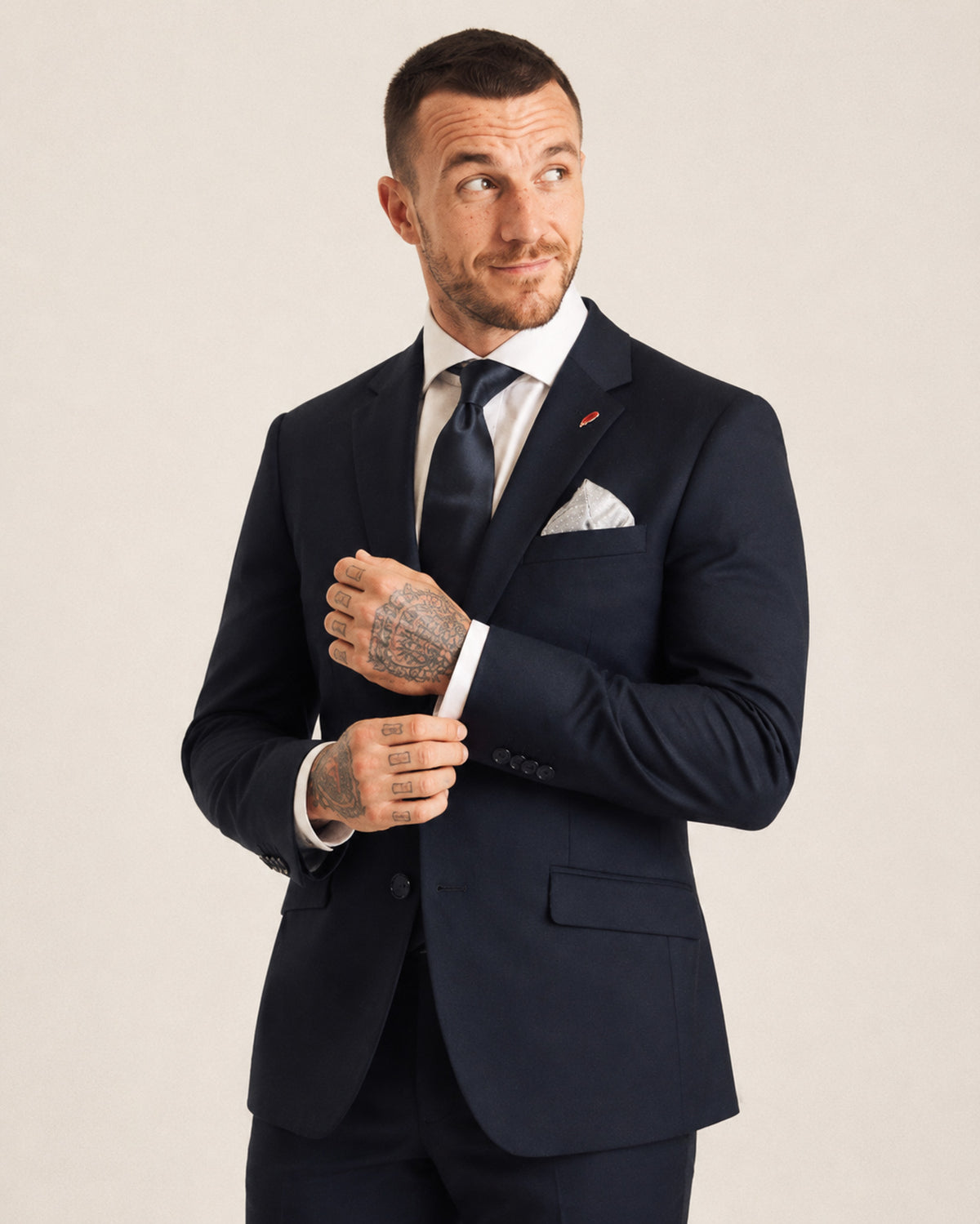 A man wearing a dark navy blue suit, white dress shirt, and navy tie, adjusting his jacket collar against a light beige studio background.