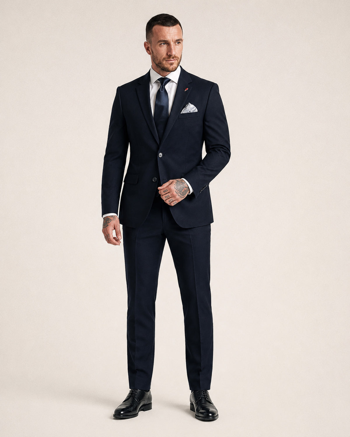 A man modeling a dark navy blue two-piece suit with a white dress shirt, a navy patterned tie, a white pocket square, and black dress shoes, standing against a solid light beige background.