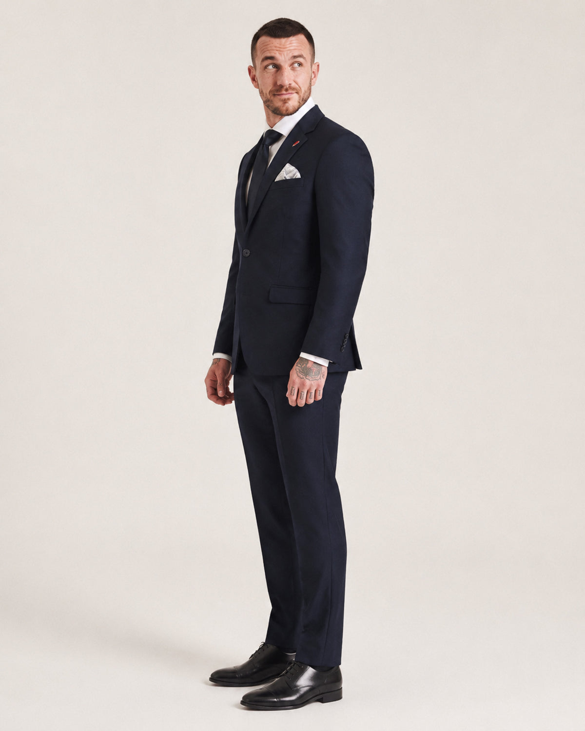 A man modeling a dark navy blue two-piece suit with a white dress shirt, black tie, and a light-colored pocket square, standing against a plain light beige studio background.