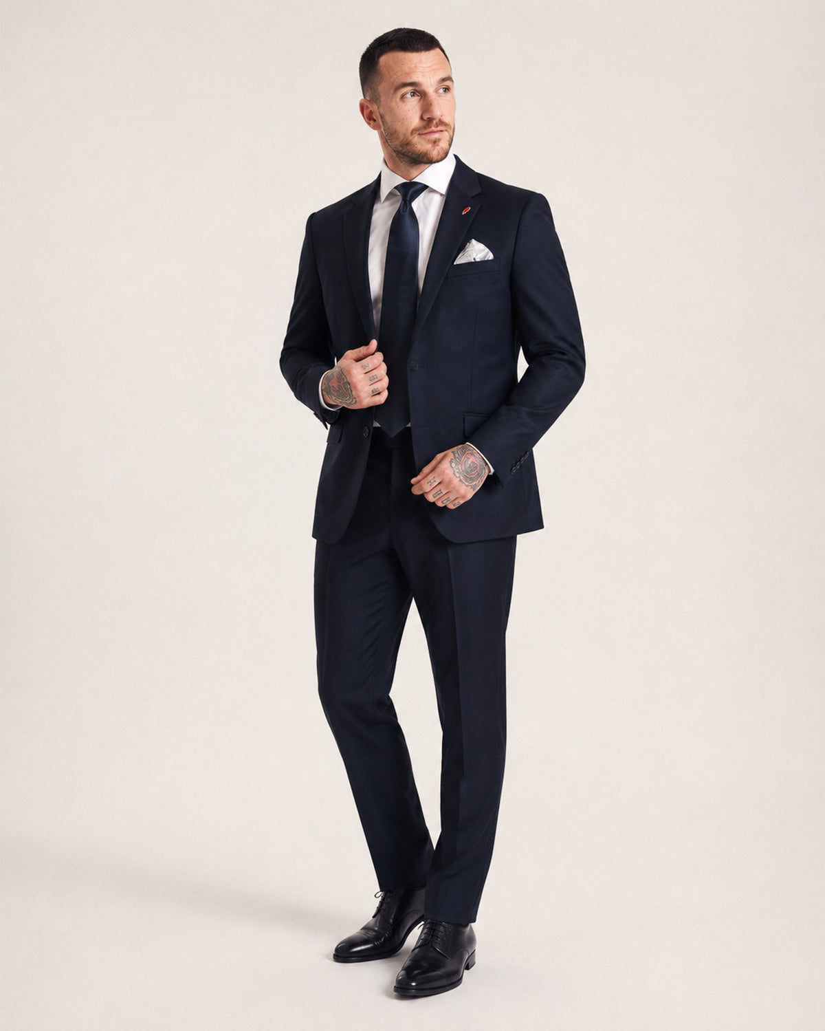 A man modeling a dark navy blue suit with a white dress shirt, a dark tie, and a white pocket square, standing against a plain light beige studio background.