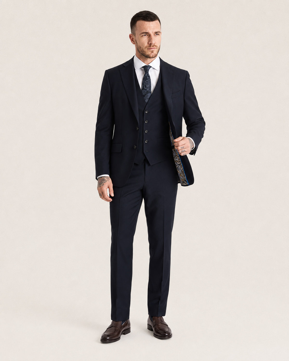 A man modeling a dark navy three-piece suit with a white dress shirt, a patterned navy tie, and dark brown dress shoes, standing against a solid light beige background.