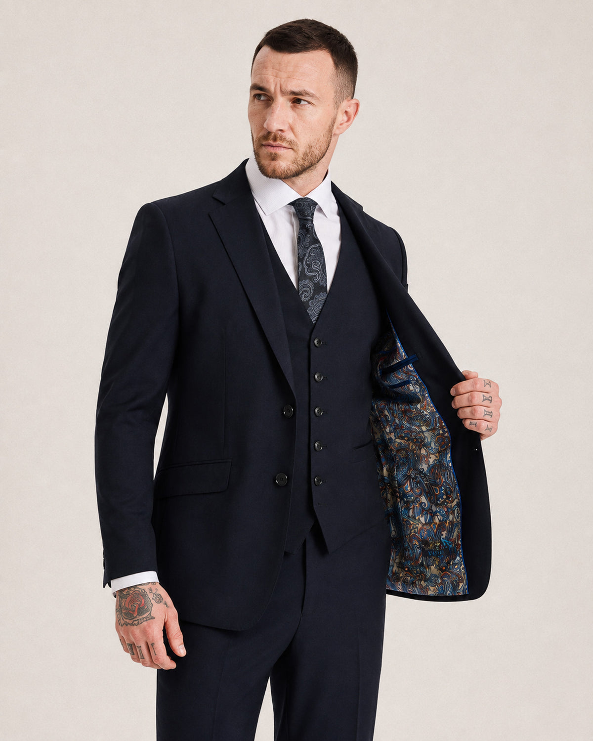 A man wearing a dark navy three-piece suit with a white dress shirt, patterned tie, and holding open the lapel to show a blue and gold patterned suit lining, against a light beige background.