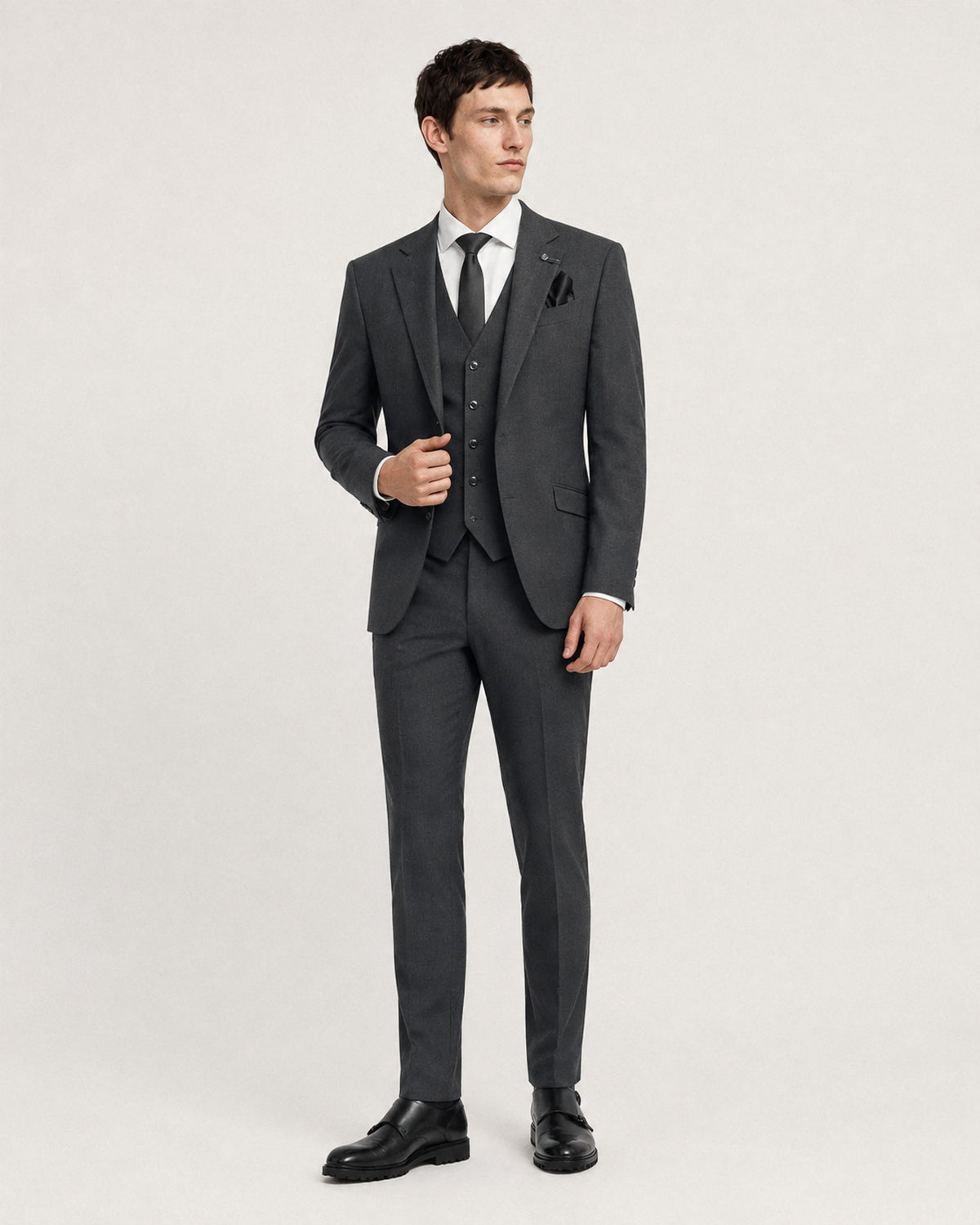A man modeling a dark gray three-piece suit with a white dress shirt, black tie, and black dress shoes against a light gray studio background.