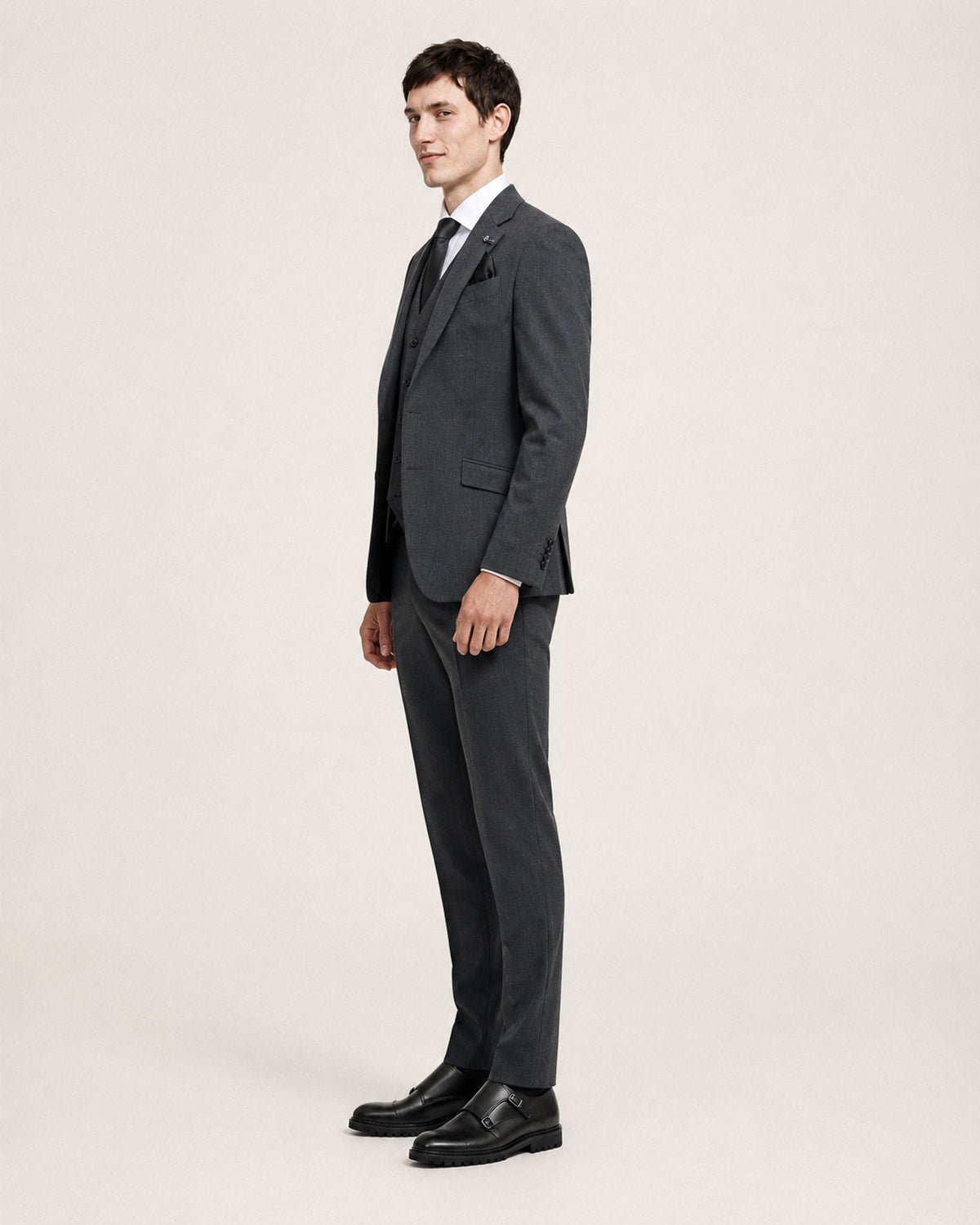 A man modeling a dark gray two-piece suit with a white dress shirt, black tie, and black dress shoes, standing in profile against a light gray studio background.