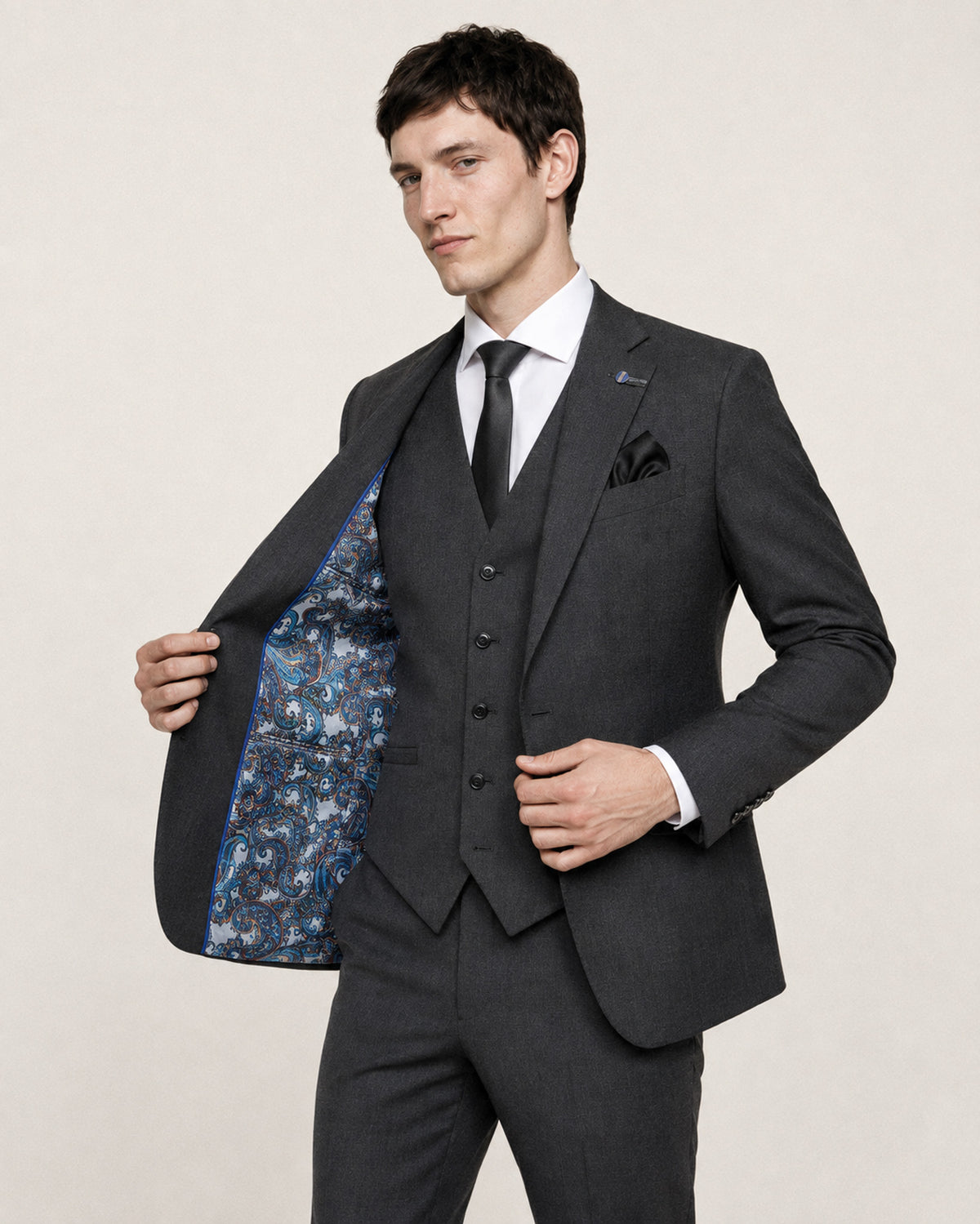 A man wearing a dark gray three-piece suit with a white dress shirt, black tie, and patterned blue lining on the jacket, standing against a light beige background.