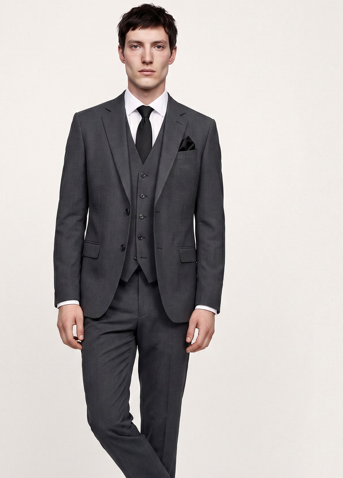 A man modeling a dark gray three-piece suit with a white dress shirt, black tie, and black pocket square against a light beige studio background.