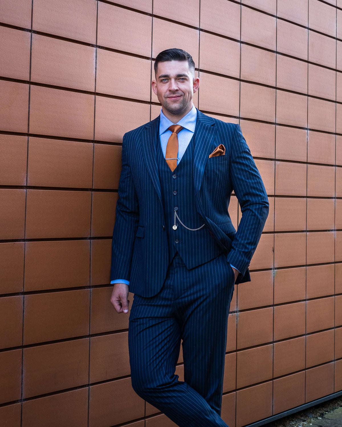 A man wearing a dark blue pinstripe three-piece suit with a light blue shirt, orange tie, and orange pocket square, posing against a brown tiled wall.