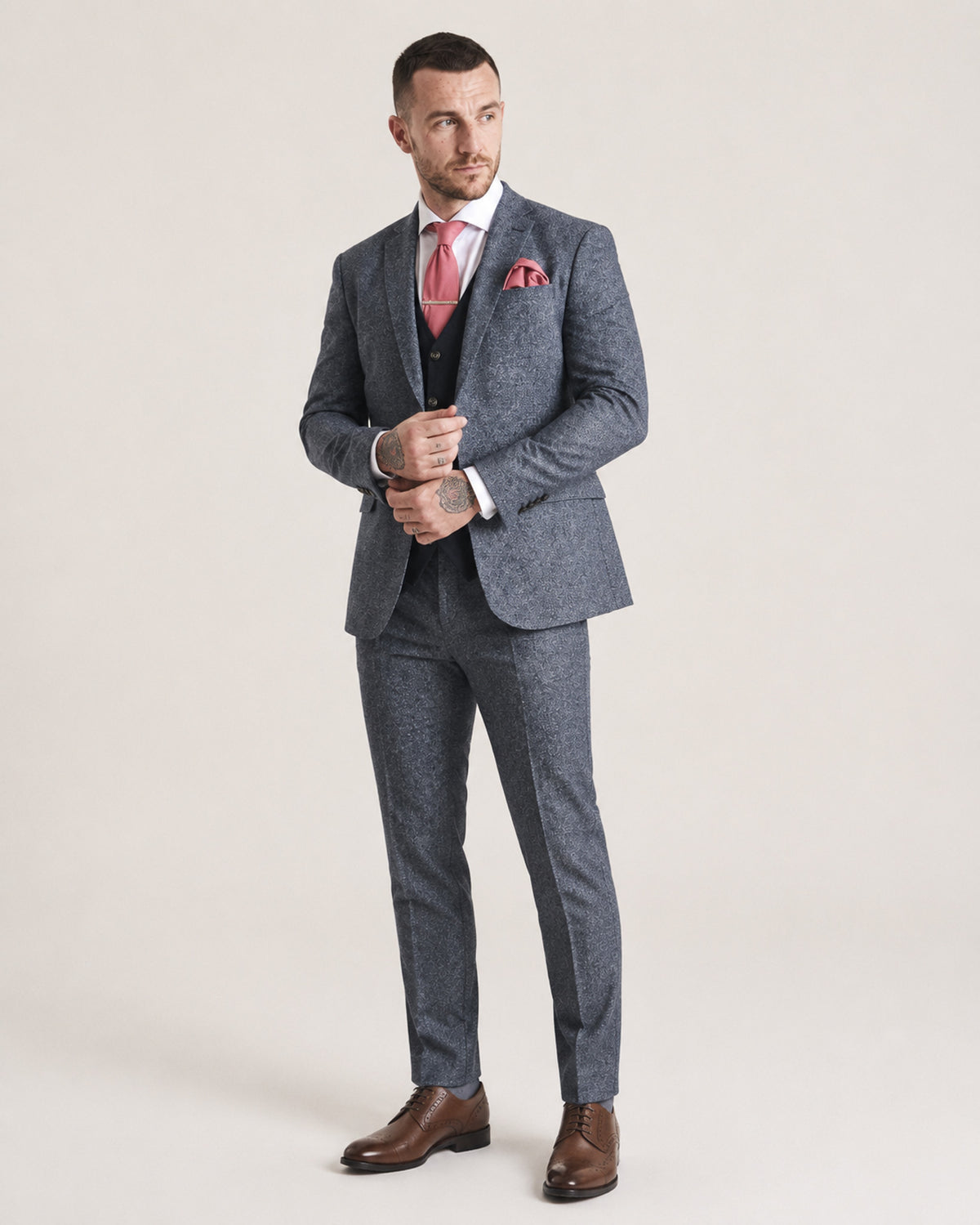 A man modeling a grey micro-houndstooth suit with a white dress shirt, a pink tie, a pink pocket square, and brown leather dress shoes, standing against a light beige studio background.