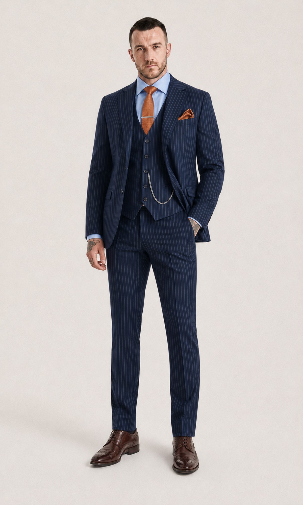 A man modeling a navy blue pinstripe three-piece suit with a light blue dress shirt, orange tie, orange pocket square, and brown leather dress shoes, standing against a light beige studio background.
