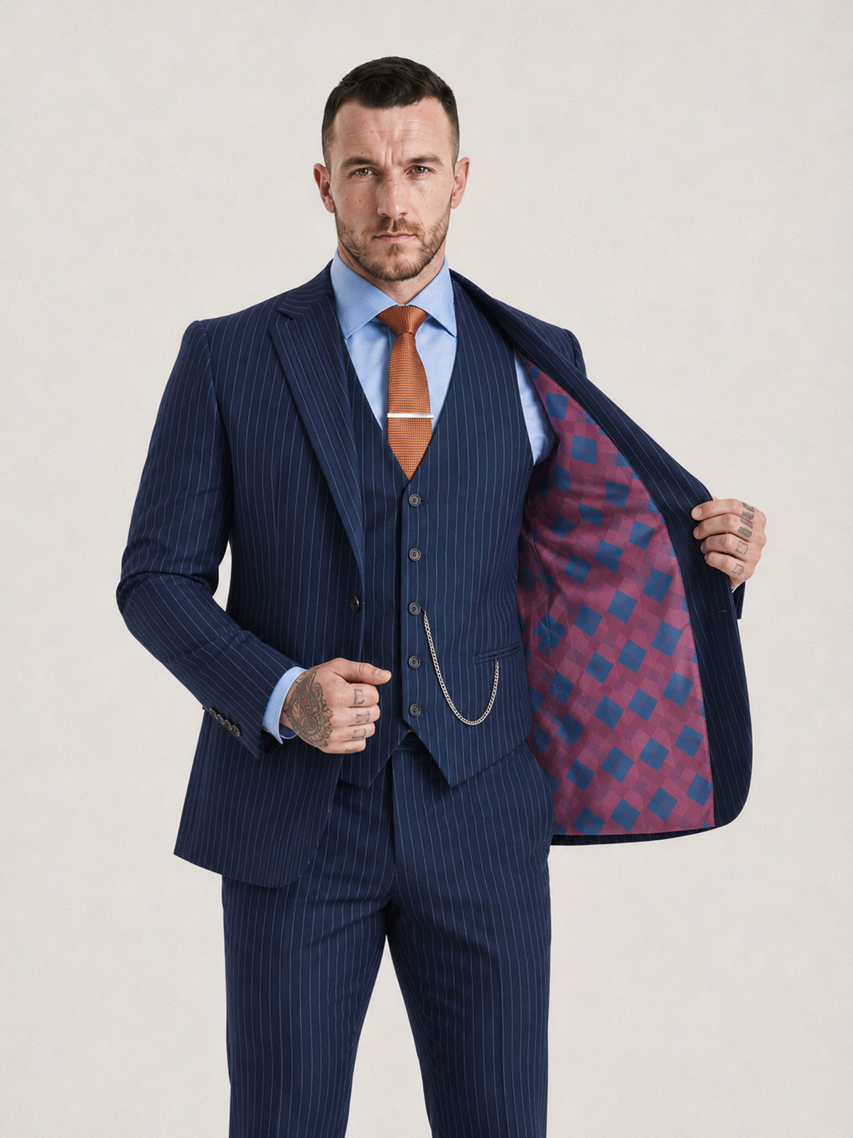 A man modeling a navy blue pinstripe three-piece suit, including a jacket, vest, and trousers, styled with a light blue dress shirt, orange tie, and silver chain. He is holding open the jacket to show a red and blue argyle patterned lining, set against a plain light gray studio background.