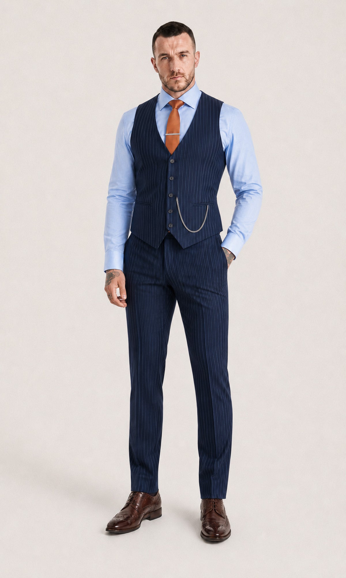 A man modeling a navy blue pinstripe three-piece suit, consisting of a vest, trousers, and matching trousers, styled with a light blue dress shirt, orange tie, and brown leather dress shoes against a light beige studio background.