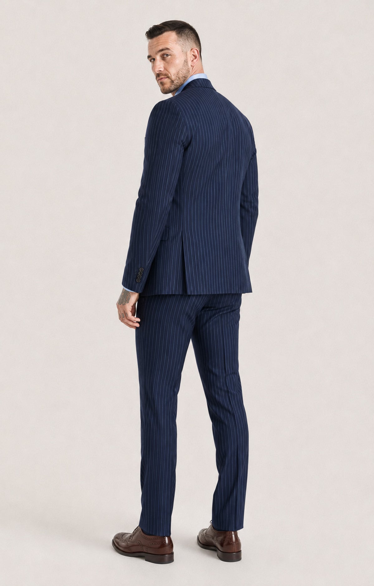 A man wearing a navy blue pinstripe suit, shown from a three-quarters back view, standing against a light beige studio background.