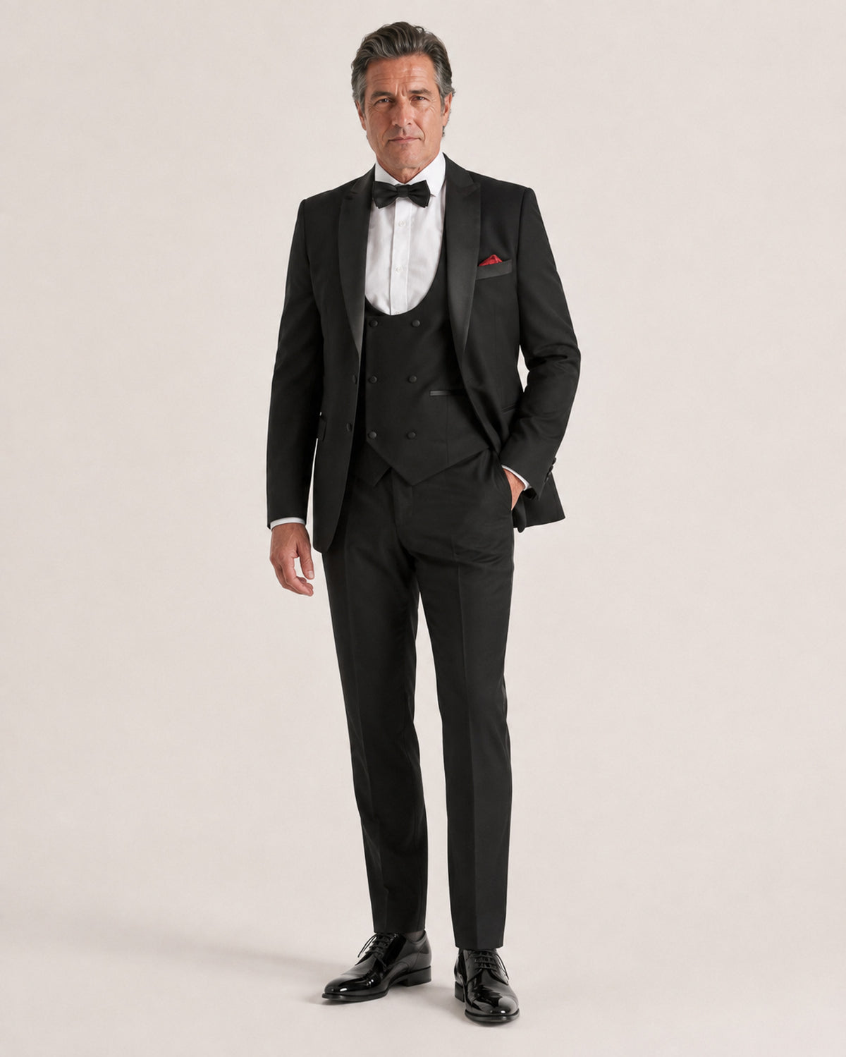 A man modeling a black three-piece tuxedo with satin lapels, a white dress shirt, a black bow tie, and a red pocket square, standing against a light beige studio background.