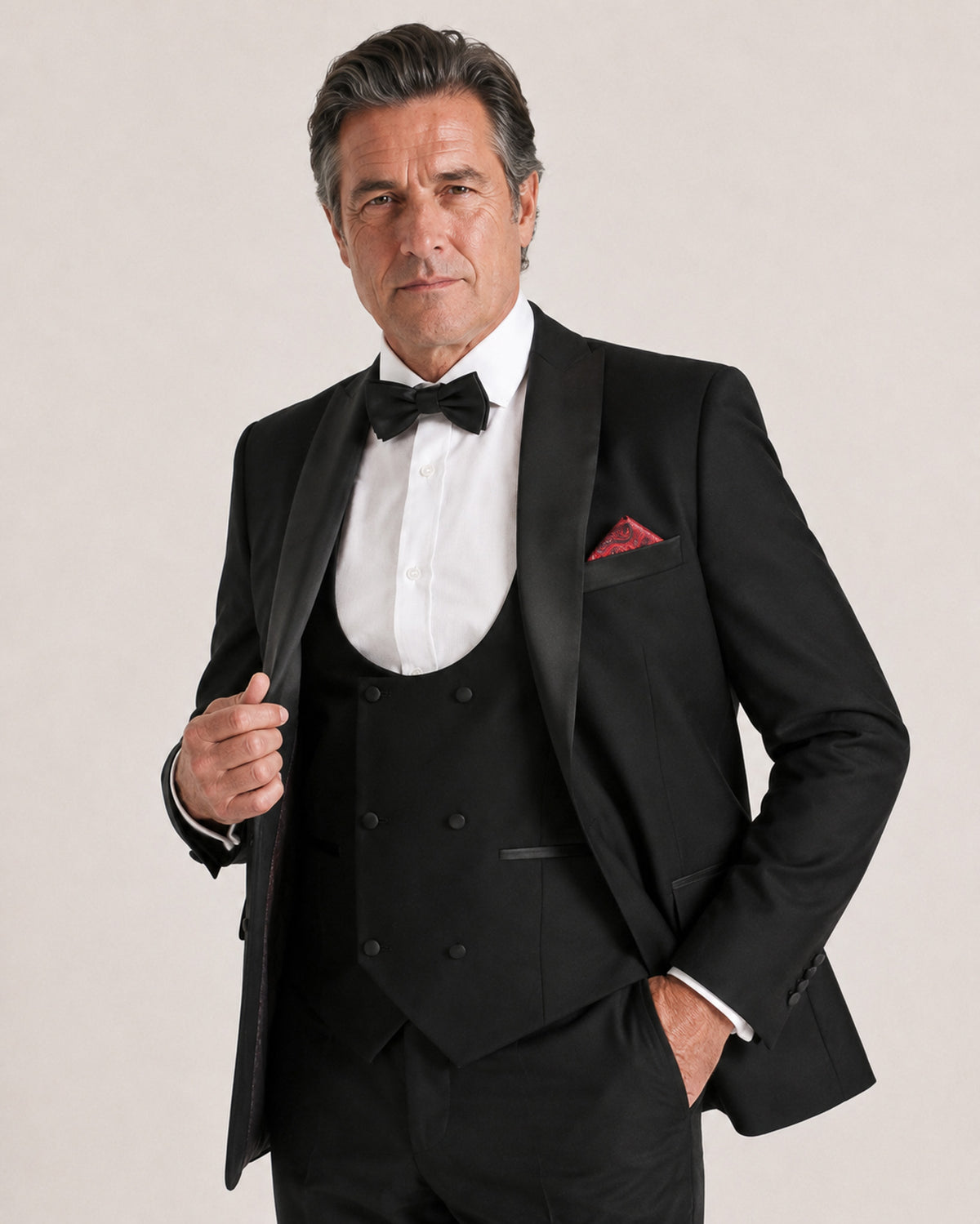 A man wearing a black tuxedo with a black vest, white dress shirt, black bow tie, and a red pocket square, posed against a light beige studio background.