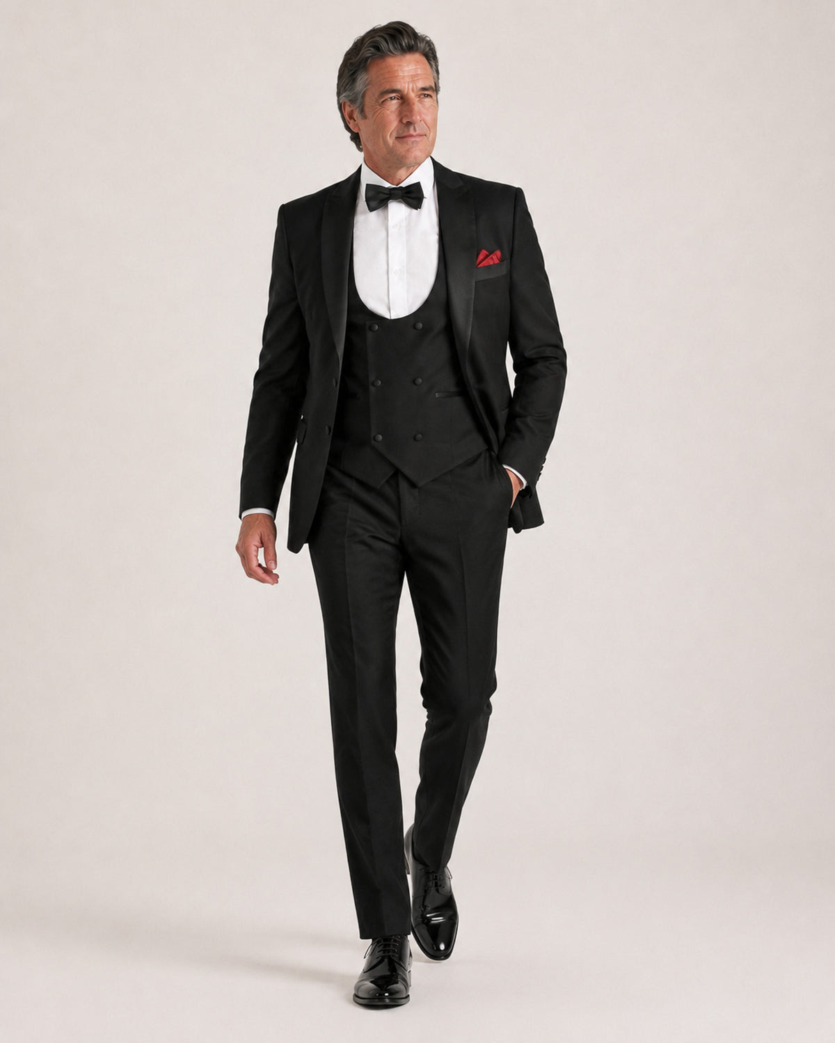 A man modeling a black tuxedo with a satin peak lapel, matching vest and trousers, a white dress shirt, a black bow tie, and a red pocket square, posed against a light beige studio background.