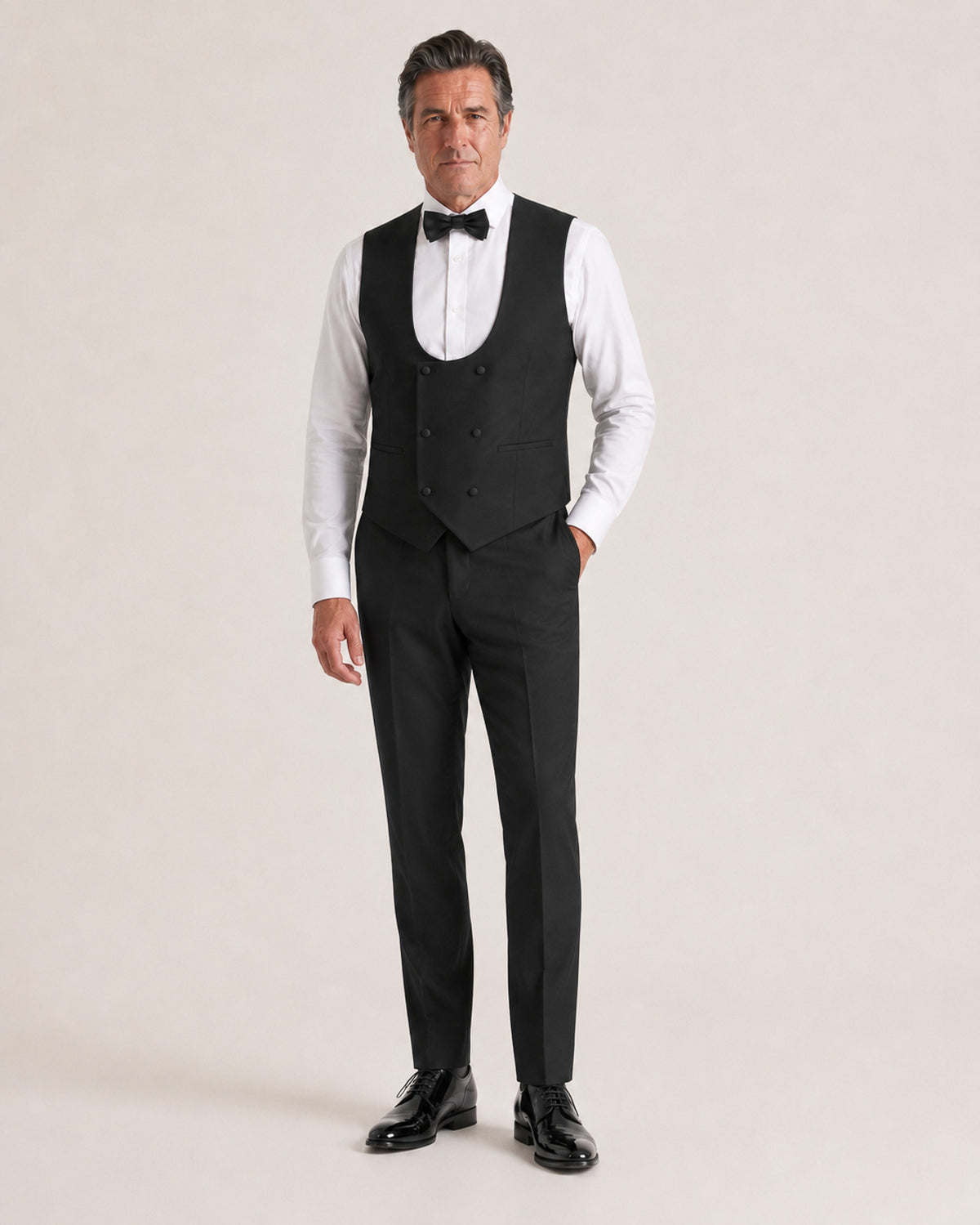 A man modeling a black formal tailoring set consisting of a waistcoat, matching trousers, a white dress shirt, and a black bow tie, standing against a solid light beige background.