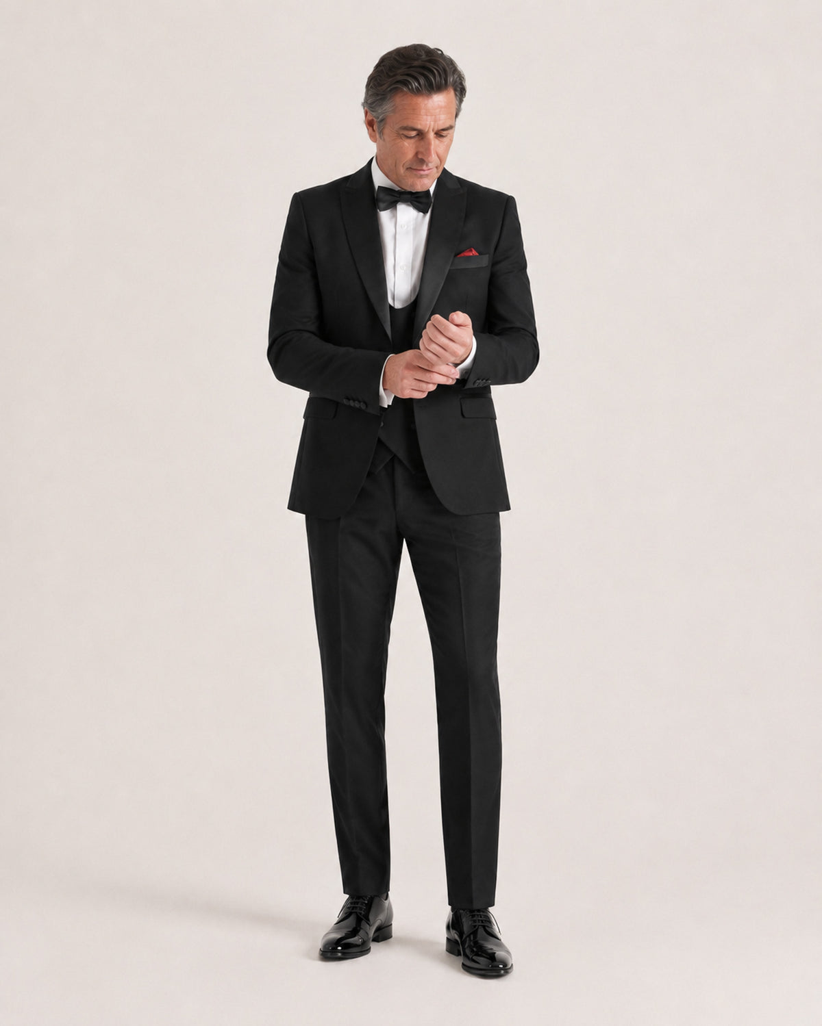 A man wearing a classic black tuxedo with black satin lapels, a white dress shirt, a black bow tie, a white pocket square, and black dress shoes, standing against a light beige studio background.
