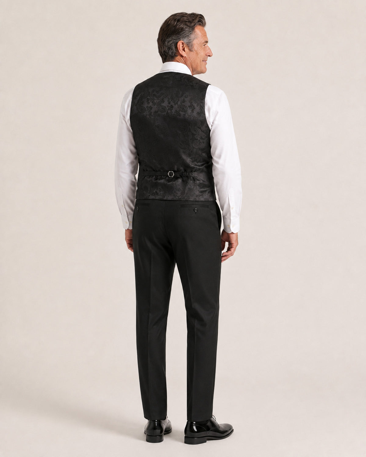 A man shown from the back wearing a black textured waistcoat over a white dress shirt, black trousers, and black dress shoes, against a plain light beige studio background.