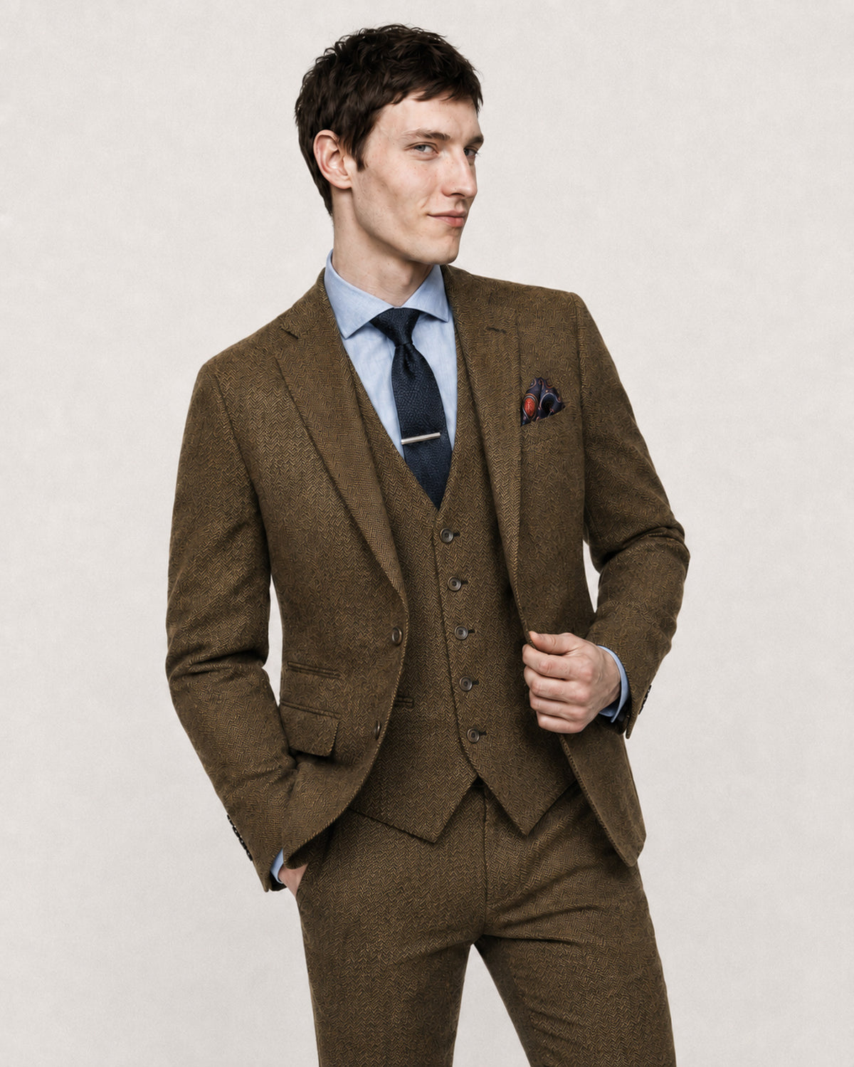 A man wearing a brown tweed three-piece suit with a matching vest and trousers, styled with a light blue dress shirt, a dark navy tie, and a patterned pocket square, against a light gray background.