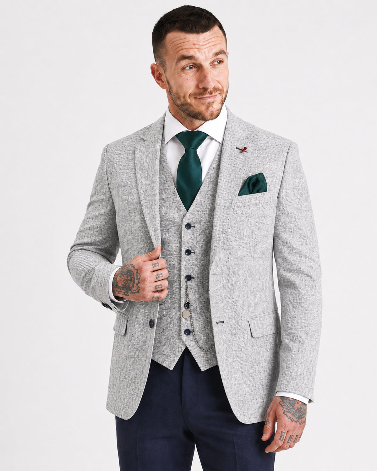 A man modeling a light gray three-piece suit with a matching vest and blazer, styled with a white dress shirt, a dark green tie, a green pocket square, and navy trousers, against a plain white studio background.