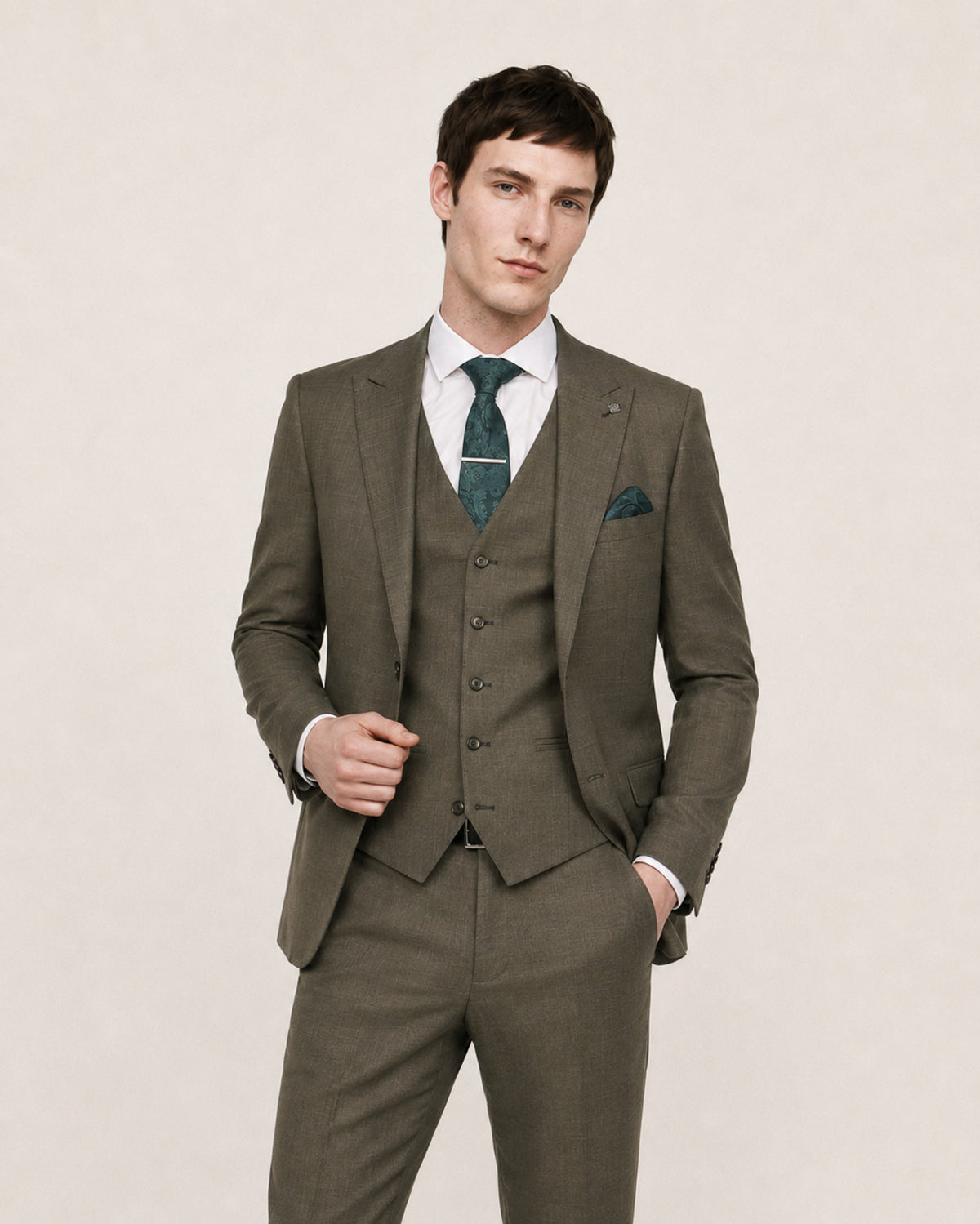 A man modeling a three-piece suit in olive green, featuring a jacket, matching vest, trousers, a white dress shirt, a patterned green tie, and a matching pocket square, set against a plain beige background.