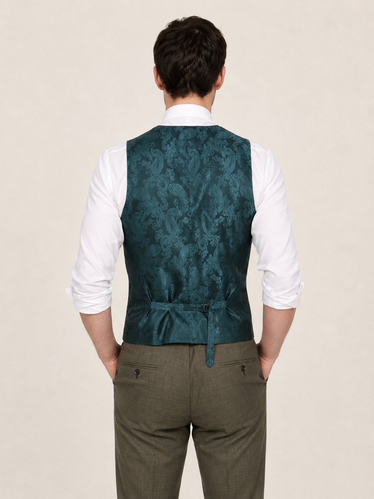 A man viewed from behind wearing a teal jacquard patterned waistcoat over a white collared shirt and olive green trousers, against a plain beige background.
