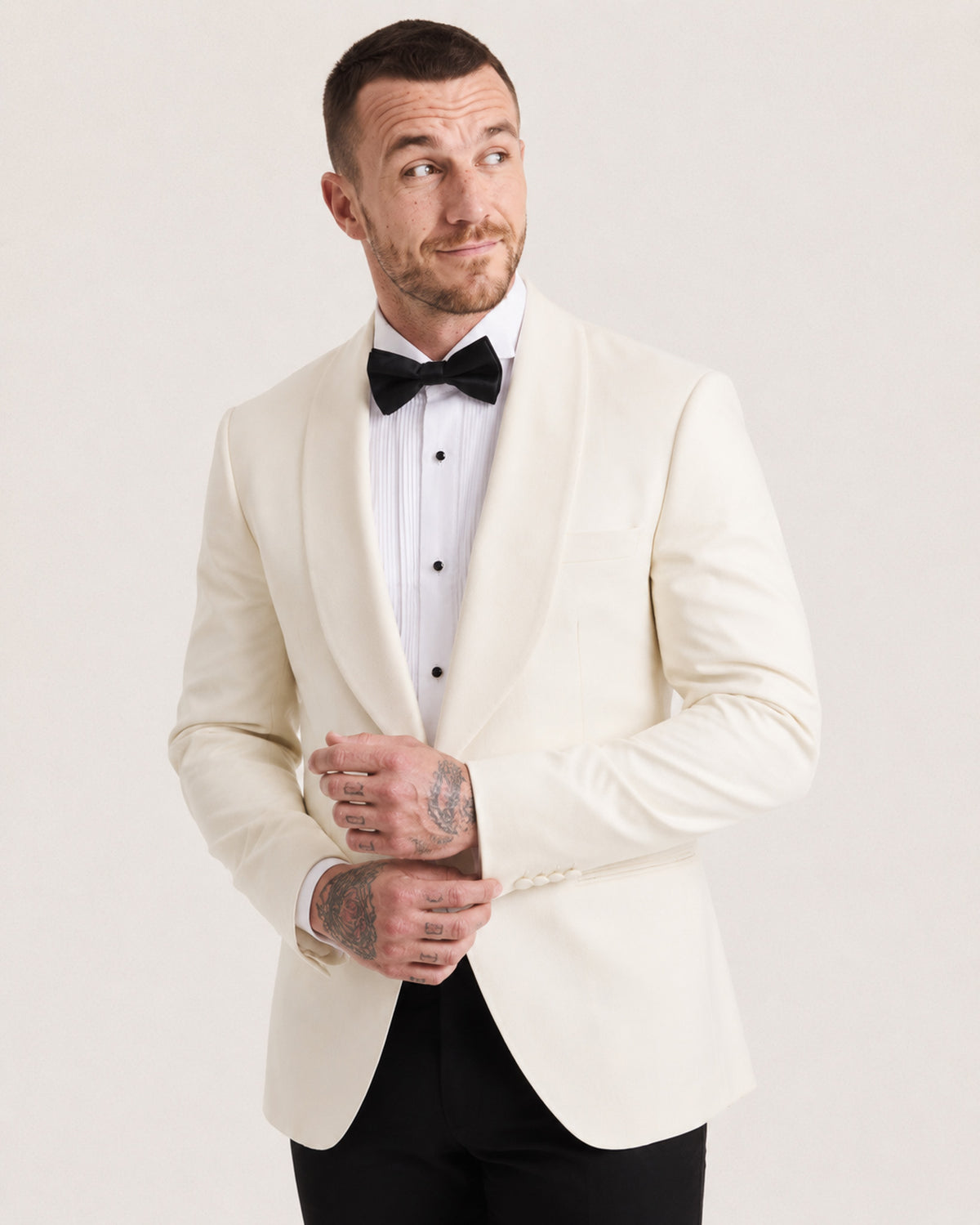 A man wearing a cream-colored shawl-lapel tuxedo jacket over a white tuxedo shirt with a black bow tie and black trousers, standing against a light gray studio background.