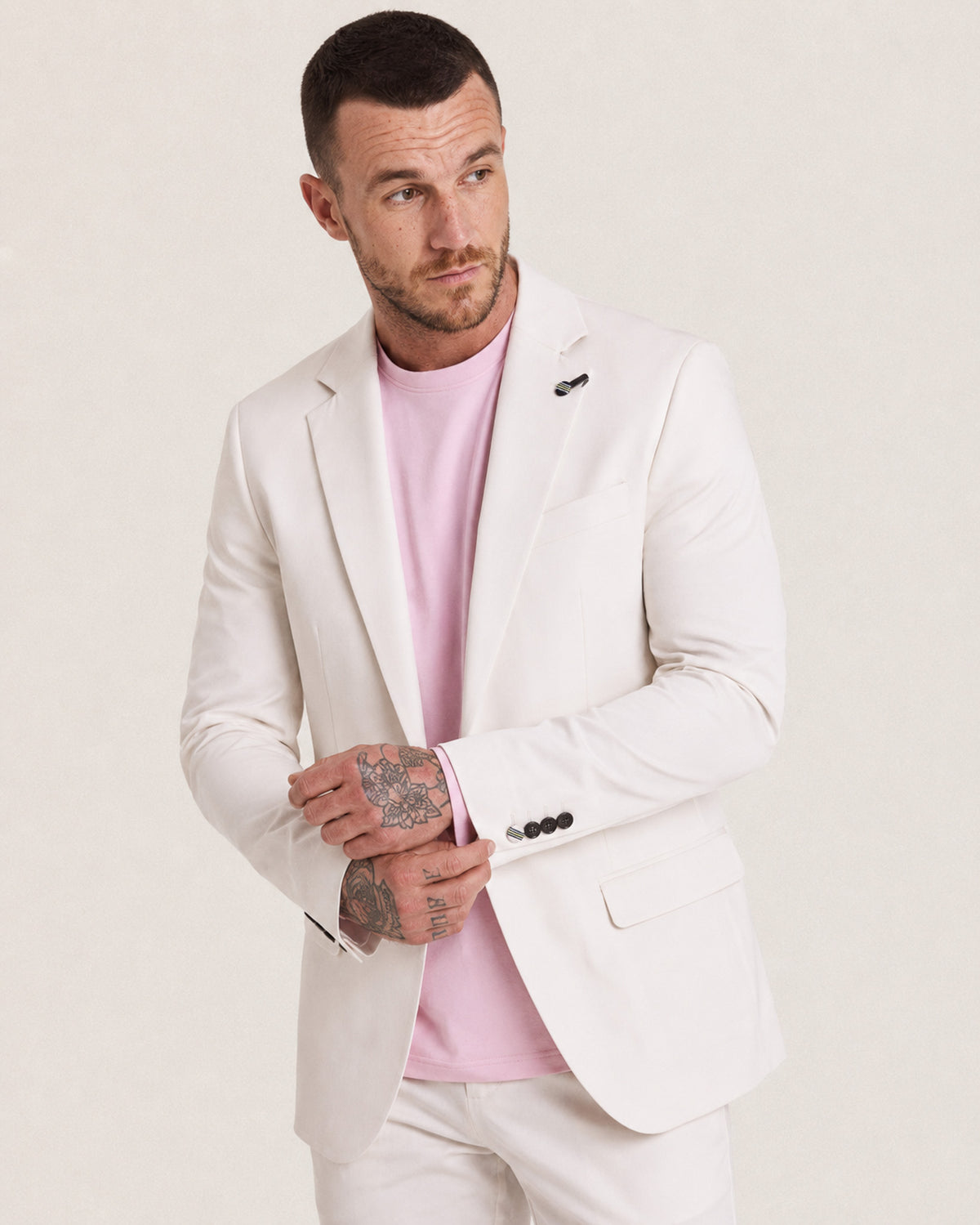 A man wearing a light cream-colored blazer and matching trousers over a light pink t-shirt, posing against a neutral off-white background.