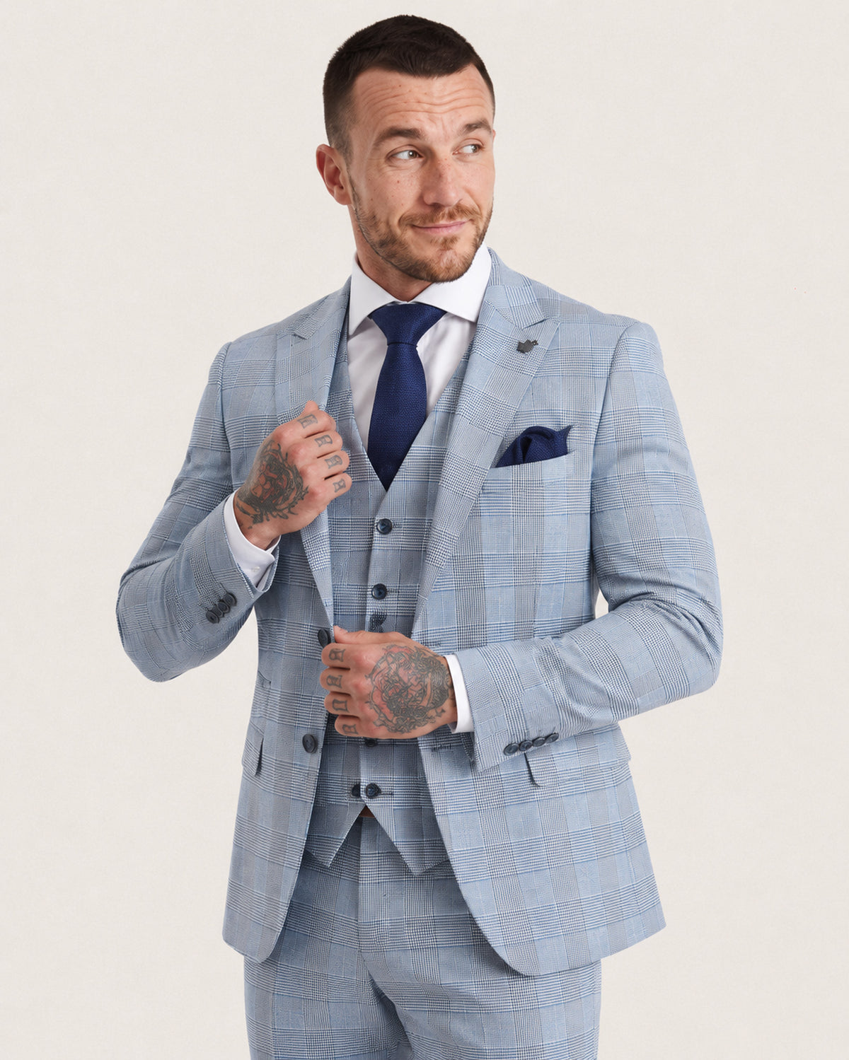 A man wearing a light blue windowpane plaid three-piece suit, white dress shirt, navy blue tie, and a matching navy pocket square, posing against a light beige studio background.