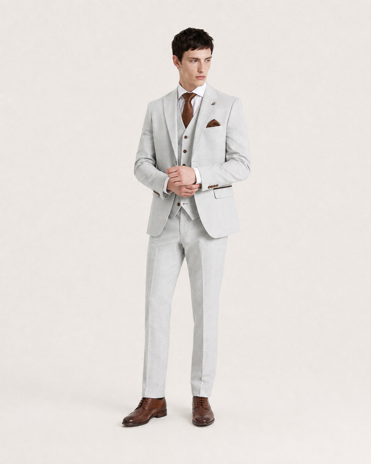 A man modeling a light gray three-piece suit with a matching vest and trousers, paired with a white shirt, brown tie, brown pocket square, and brown dress shoes, against a plain off-white studio background.