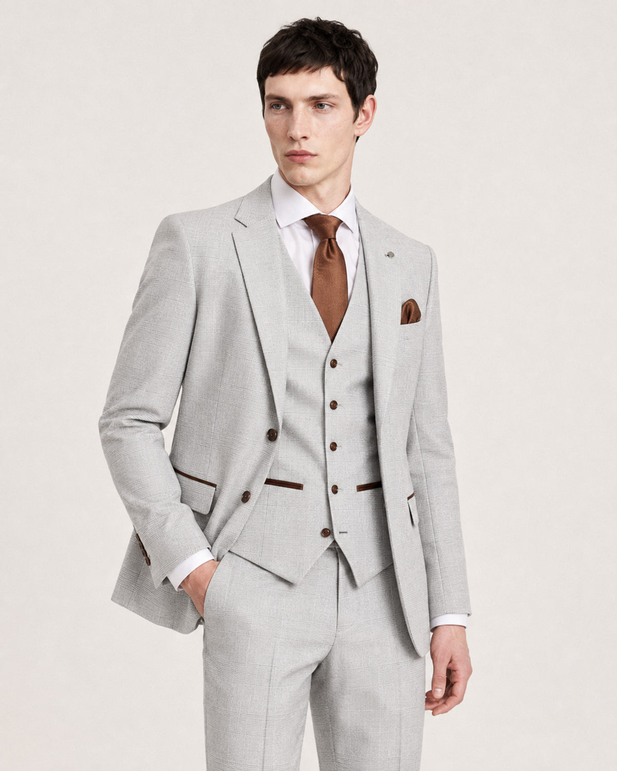 A man modeling a light gray three-piece suit with brown trim, a white dress shirt, a brown tie, and a matching pocket square, photographed against a plain light background.