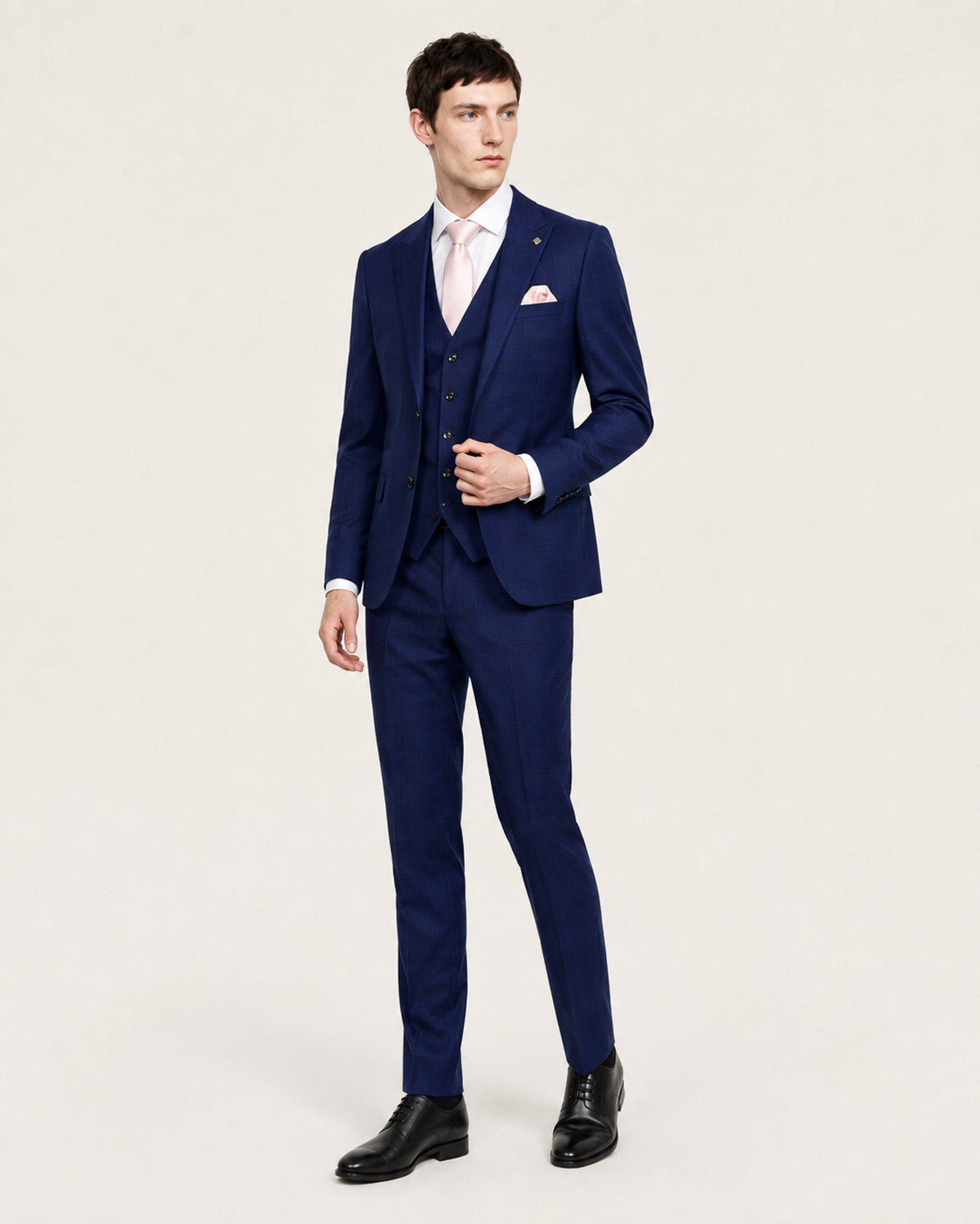 A man modeling a navy blue three-piece suit with a matching vest, white dress shirt, light pink tie, and a white pocket square, paired with black dress shoes against a plain light background.