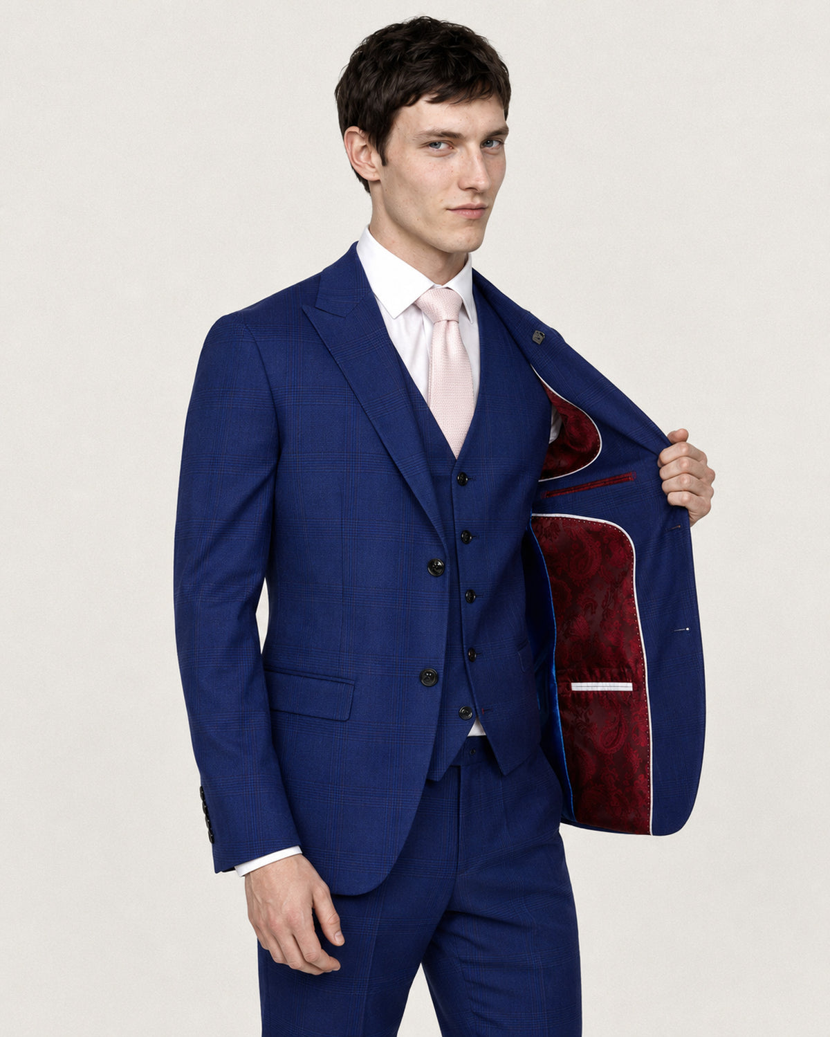 A man wearing a blue three-piece suit with a white dress shirt and light pink tie, holding open the lapel of his jacket to show a red patterned lining, against a plain light gray background.